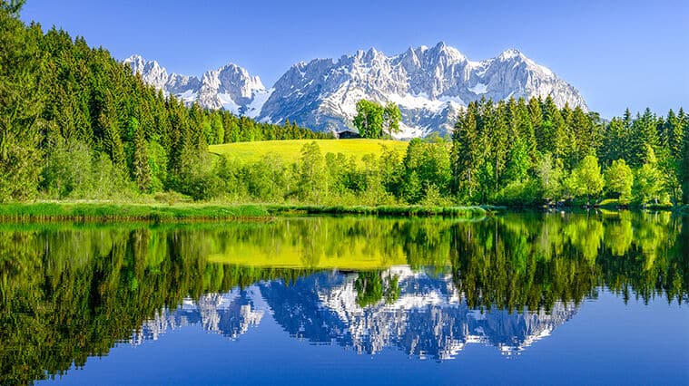 Pristine alpine lake perfectly reflecting snow-capped peaks and lush forests under a brilliant blue sky.