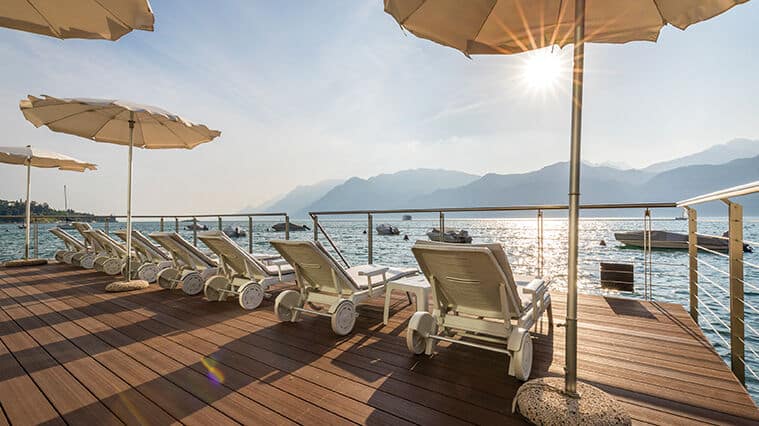 Wooden deck with white loungers and beige umbrellas overlooking a sparkling lake with mountains rising in the distance.