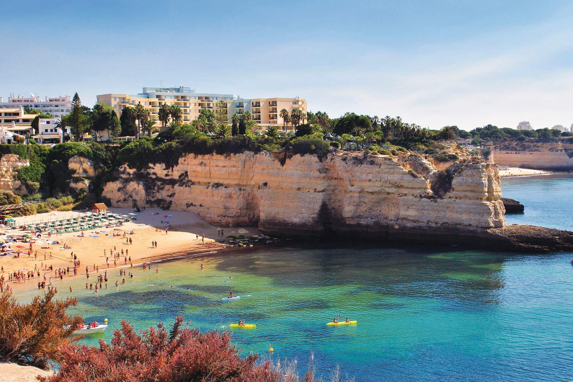 A scenic Mediterranean coastal cove with a golden sandy beach nestled beneath layered sandstone cliffs.