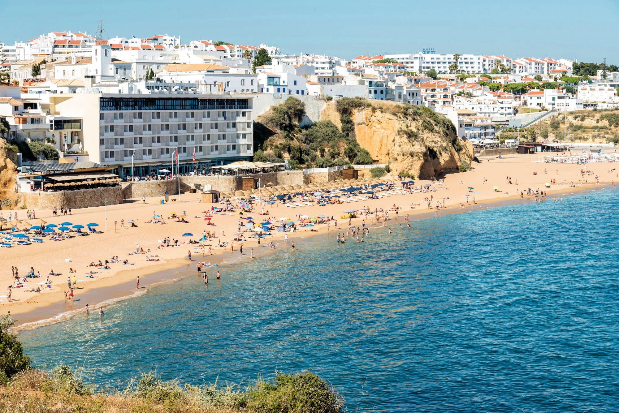 A view of a golden-sand beach with large rocky cliff, and white hillside town buildings rising in the background.