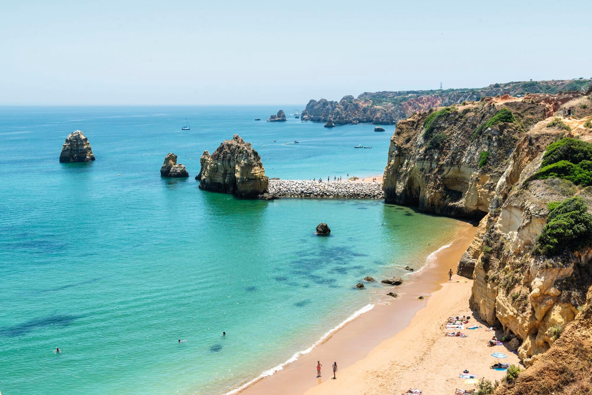 Aerial view of a Mediterranean beach with dramatic golden limestone cliffs and sea stacks rising from turquoise water.