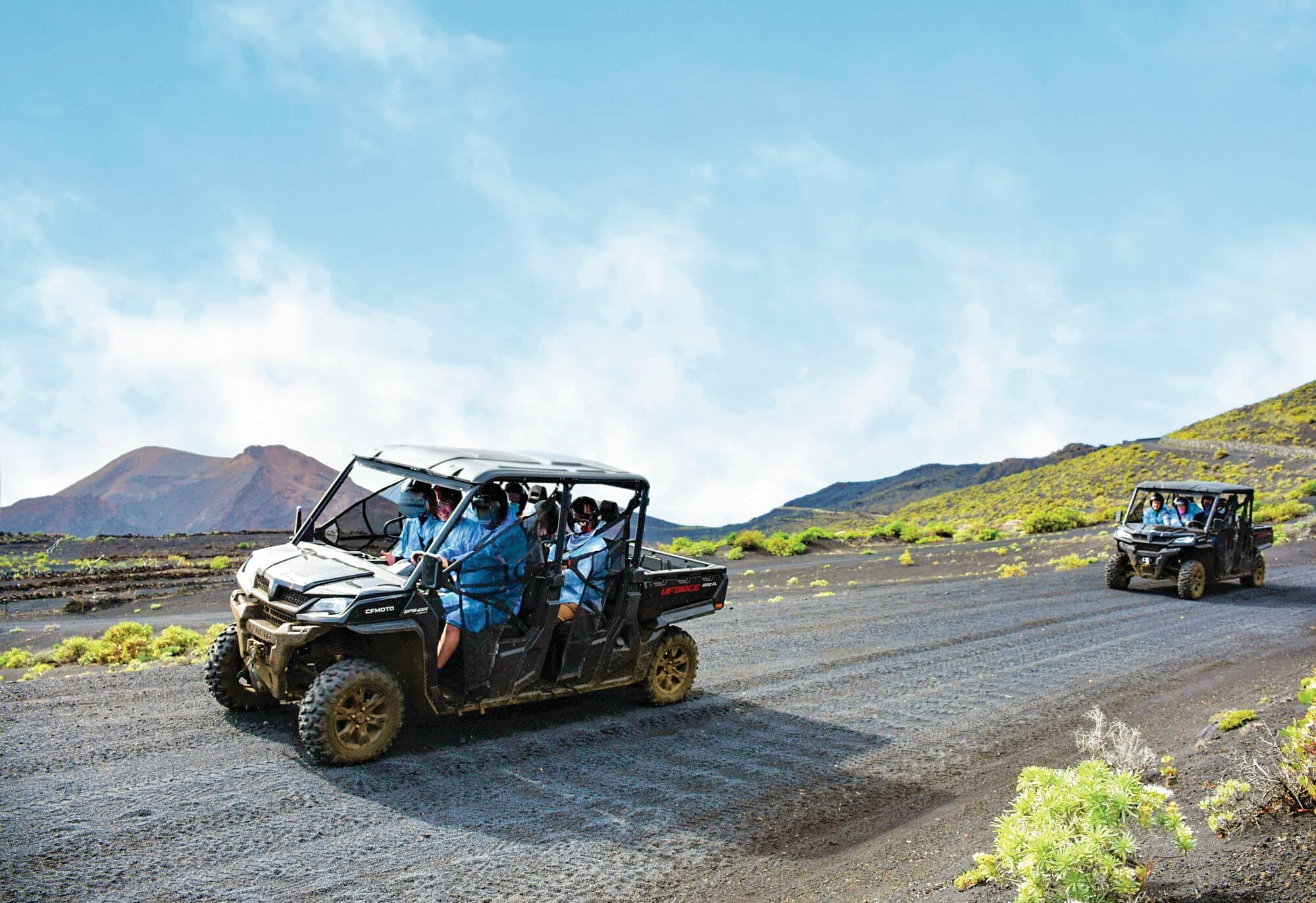 Four people in bright turquoise shirts riding a black off-road buggy on a dirt trail through a volcanic landscape.