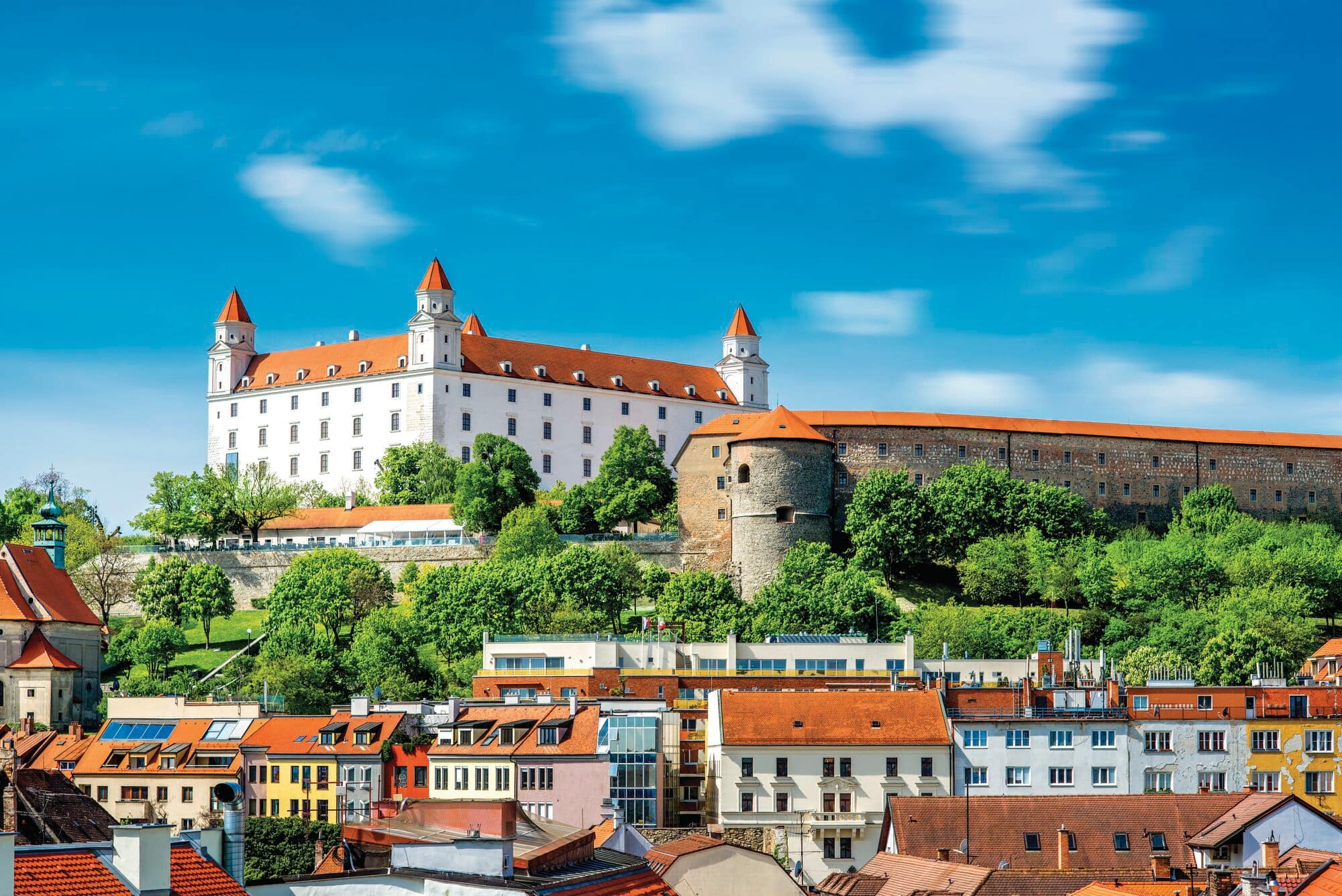 A view of Bratislava castle on a green hill ,with old houses at the bottom.