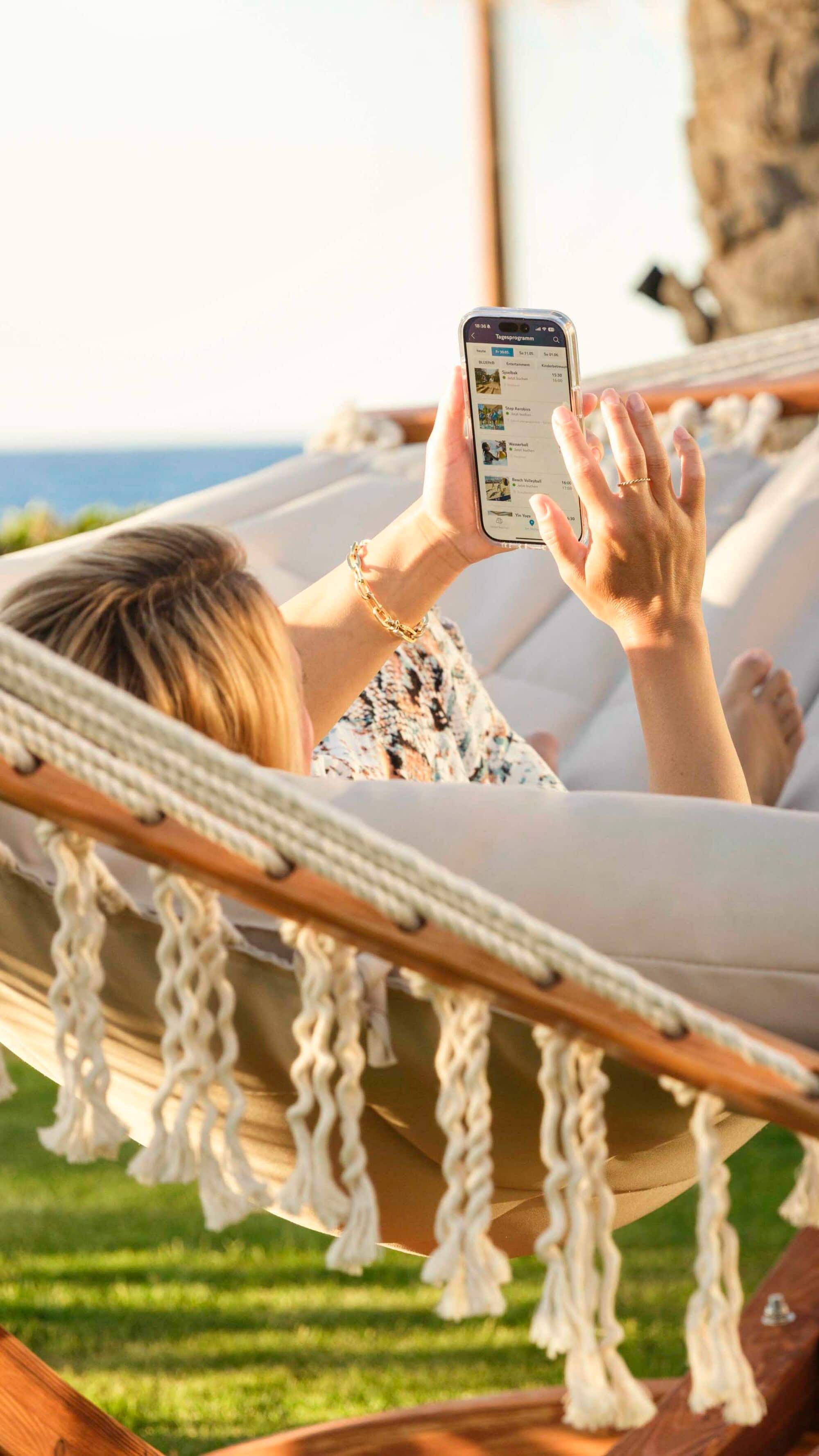 A woman laying on a hammock looking at the TUI BLUE app on her phone.