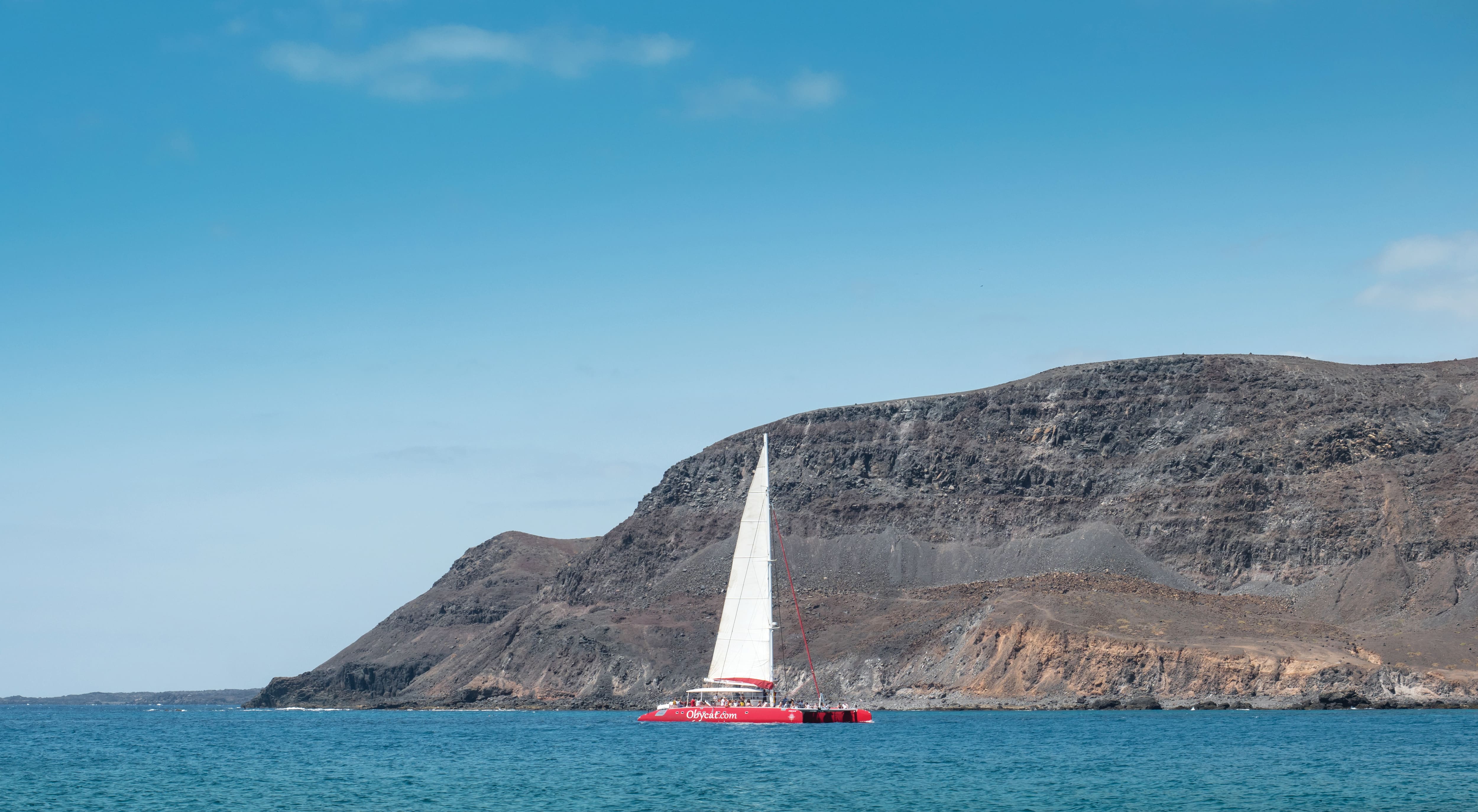 A catamaran sailing on blue water with coastal cliffs behind.
