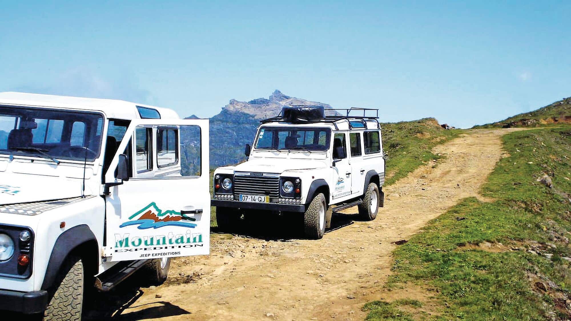 Two 4x4 expedition vehicles on a dirt track in a mountain landscape.