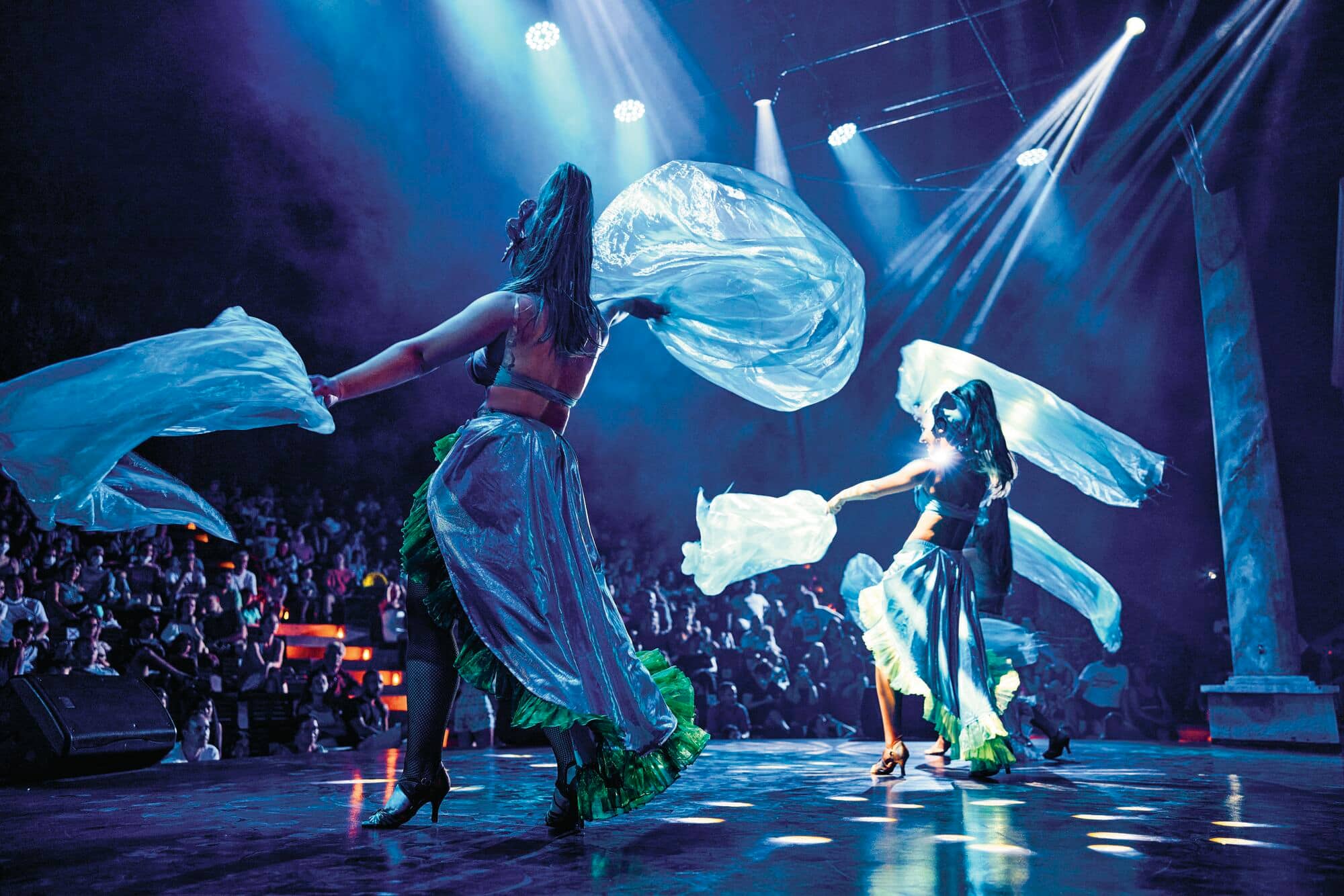 Two performers dancing with blue and green flowing fabrics under dramatic stage lighting.