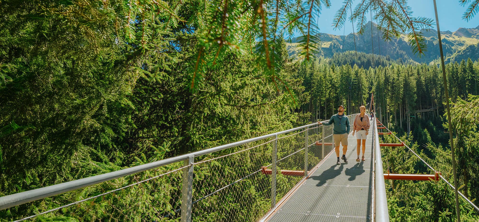 Two hikers crossing a suspension bridge through a forest canopy with mountain views in the distance on a sunny day.
