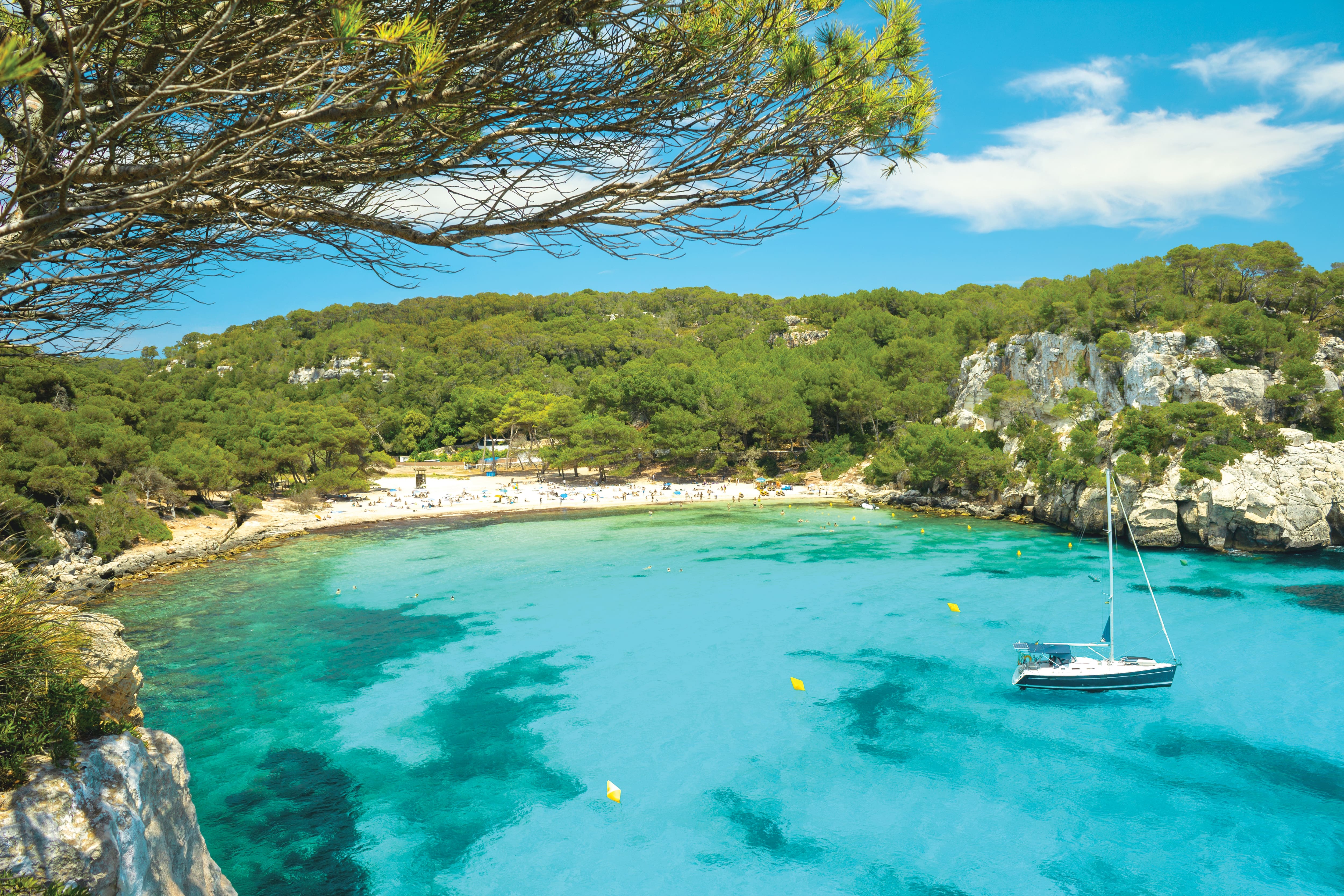 Mediterranean coastal cove with turquoise water, white sailboat, curved sandy beach with beachgoers.