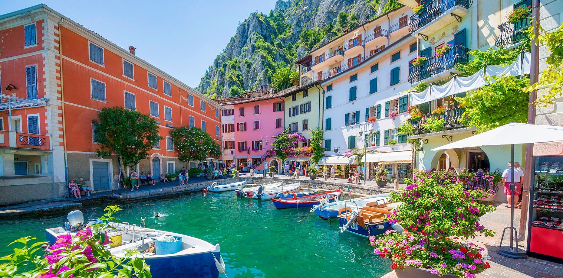  Colourful lakeside village with terracotta and pastel buildings, small boats moored in turquoise waters, backed by dramatic cliffs.