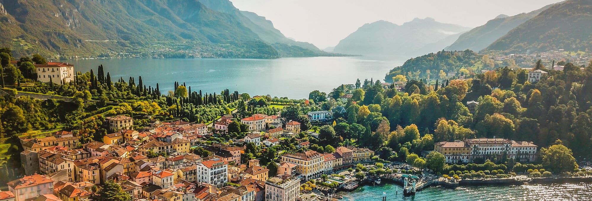 Hillside Italian village with terracotta roofs cascades toward a turquoise lake, framed by forested slopes and distant peaks.