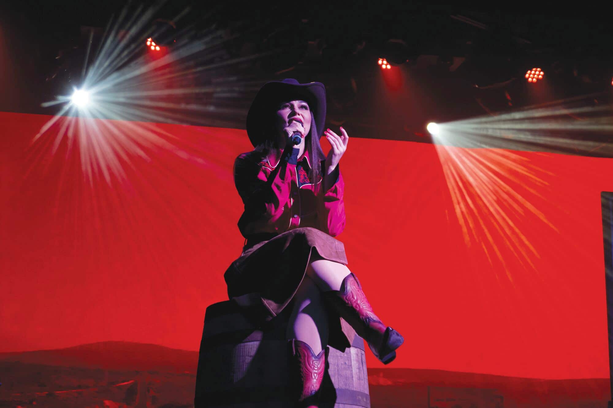 A performer in a cowboy hat sitting down and singing on stage,