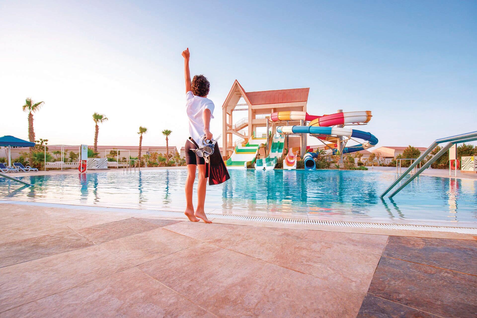 A child holding snorkelling equipment by a pool with waterslides. 