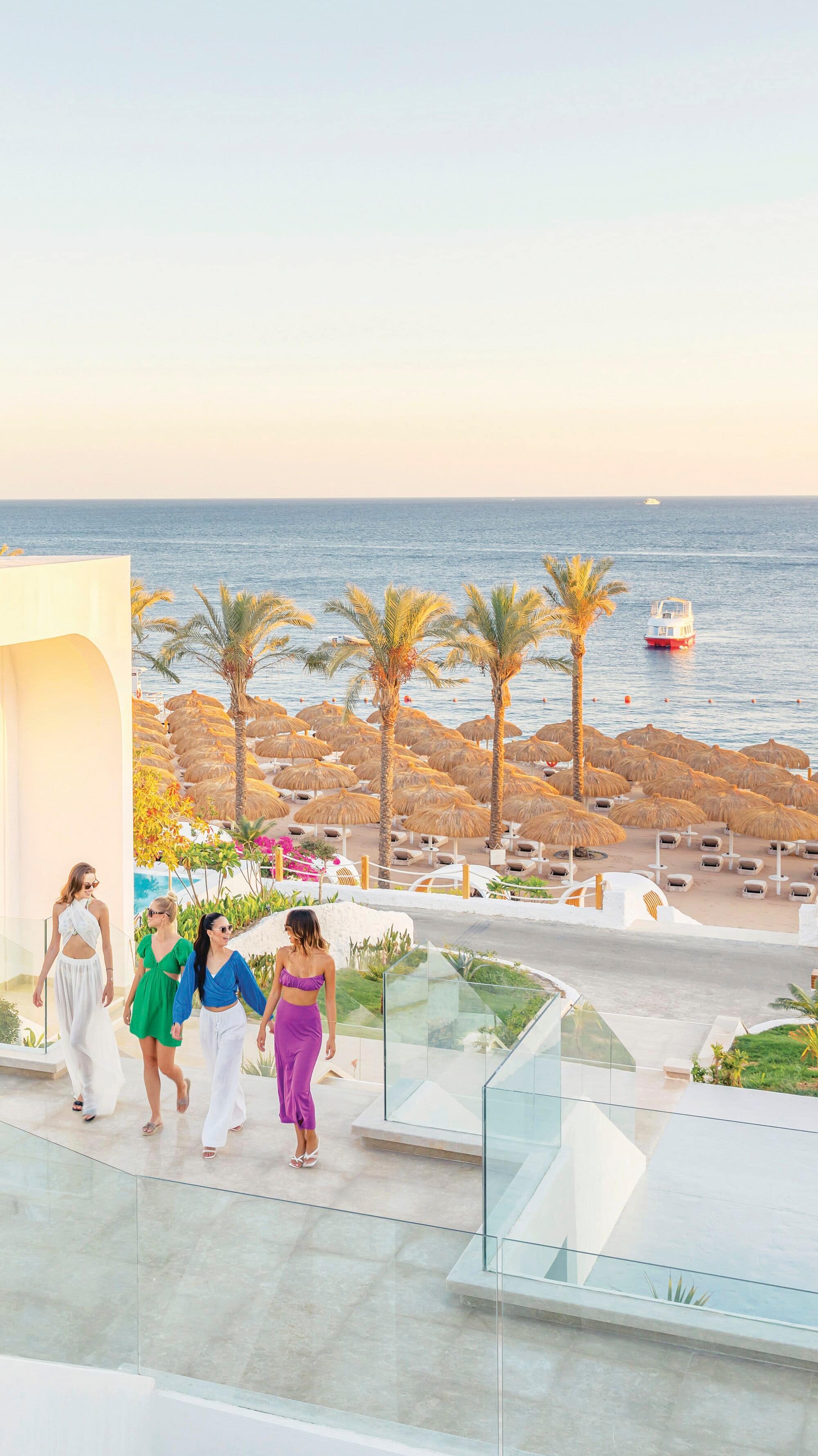  A group of friends walking up steps in the hotel grounds, with a thatched parasol-lined beach in the background. 