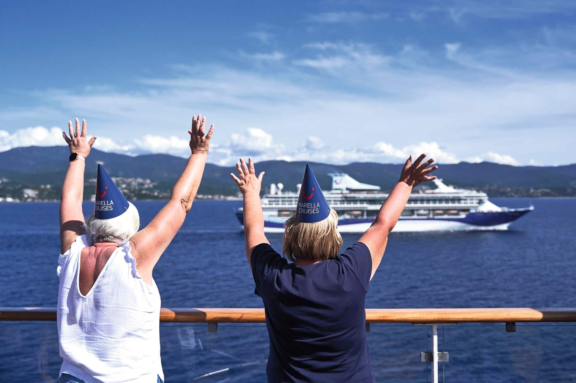 Two cruise guests wearing Marella party hats and waving at a ship.