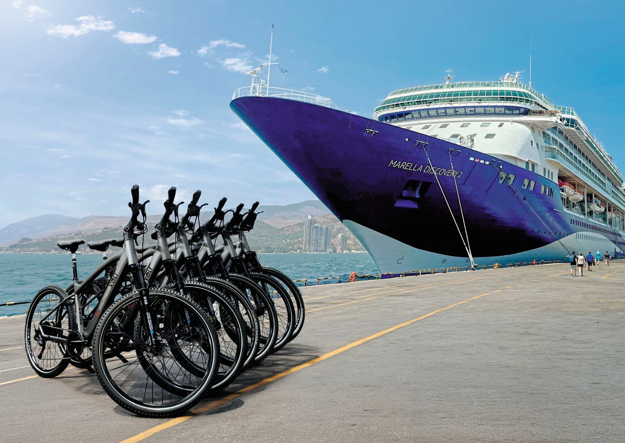 A row of e-bikes with baskets parked on a pier in front of a Marella cruise ship. Mountains are in the background.