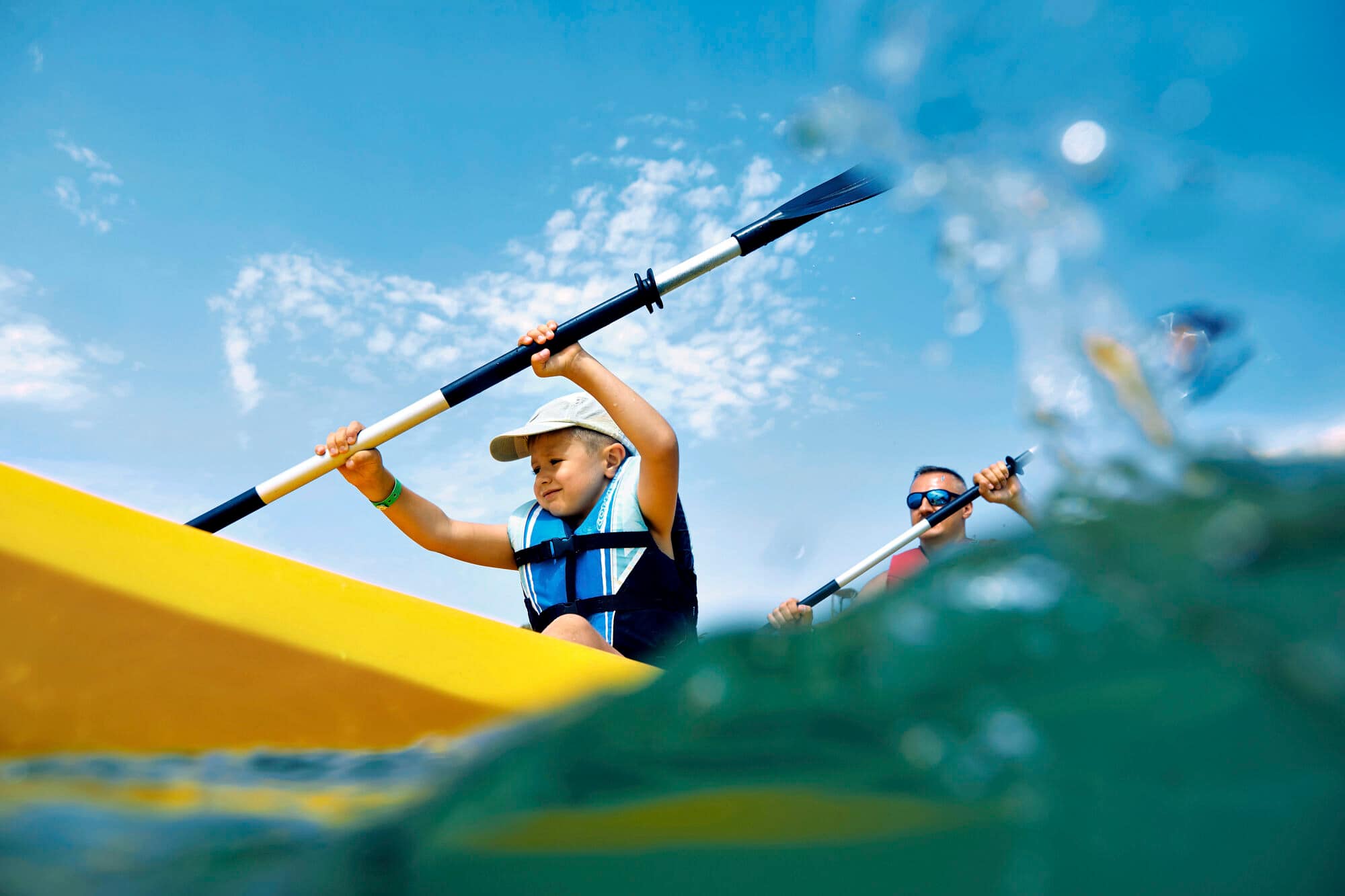 Two kayakers paddling on teal-green water under a bright blue sky with scattered clouds, photographed from water level. 
