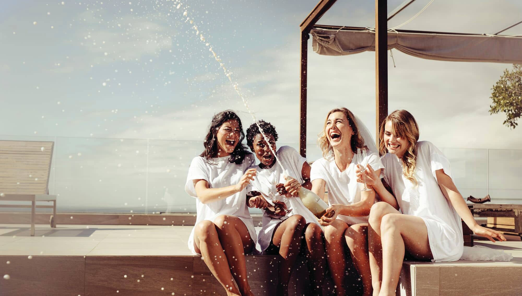 Four women in white outfits laughing together on outdoor terrace seating as confetti falls around them.
