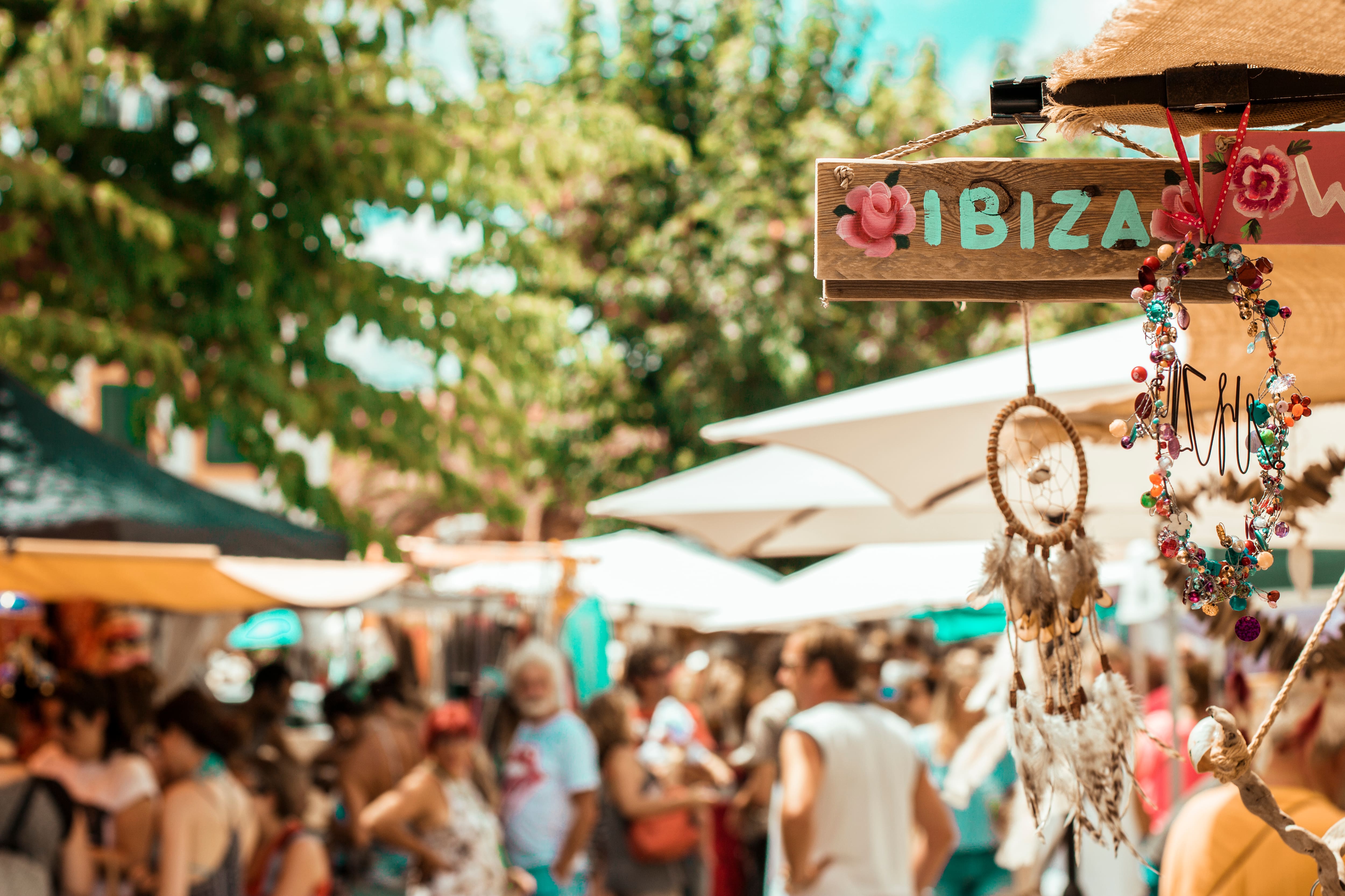 Wooden Ibiza sign and hanging craft decorations at an outdoor market with people and stalls in the background.