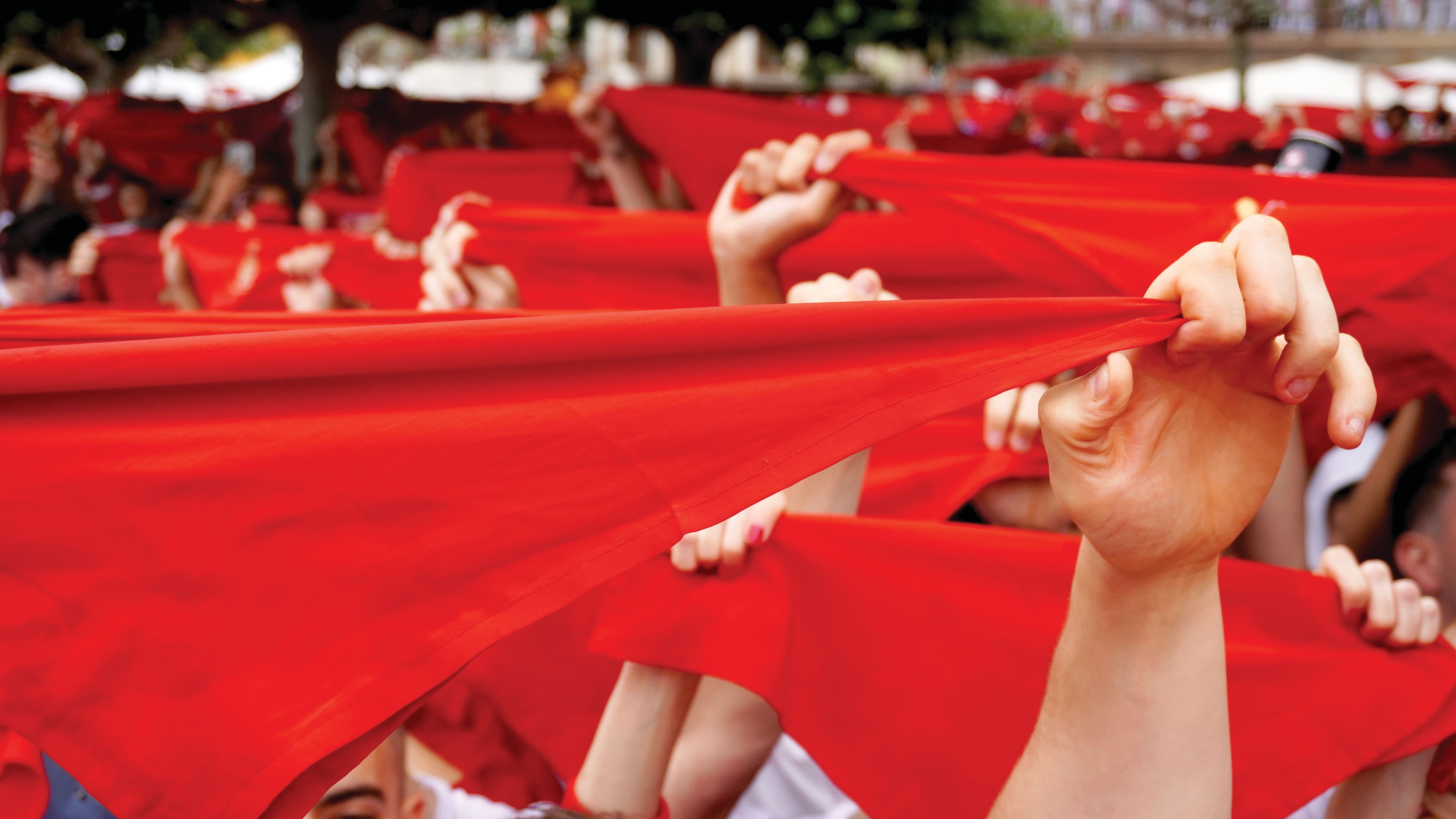 Participants at the San Fermín Festival hold bright red scarves stretched above their heads during a crowded outdoor celebration. The vivid red fabric fills the scene, creating a striking display associated with the famous festival in Pamplona, Spain.