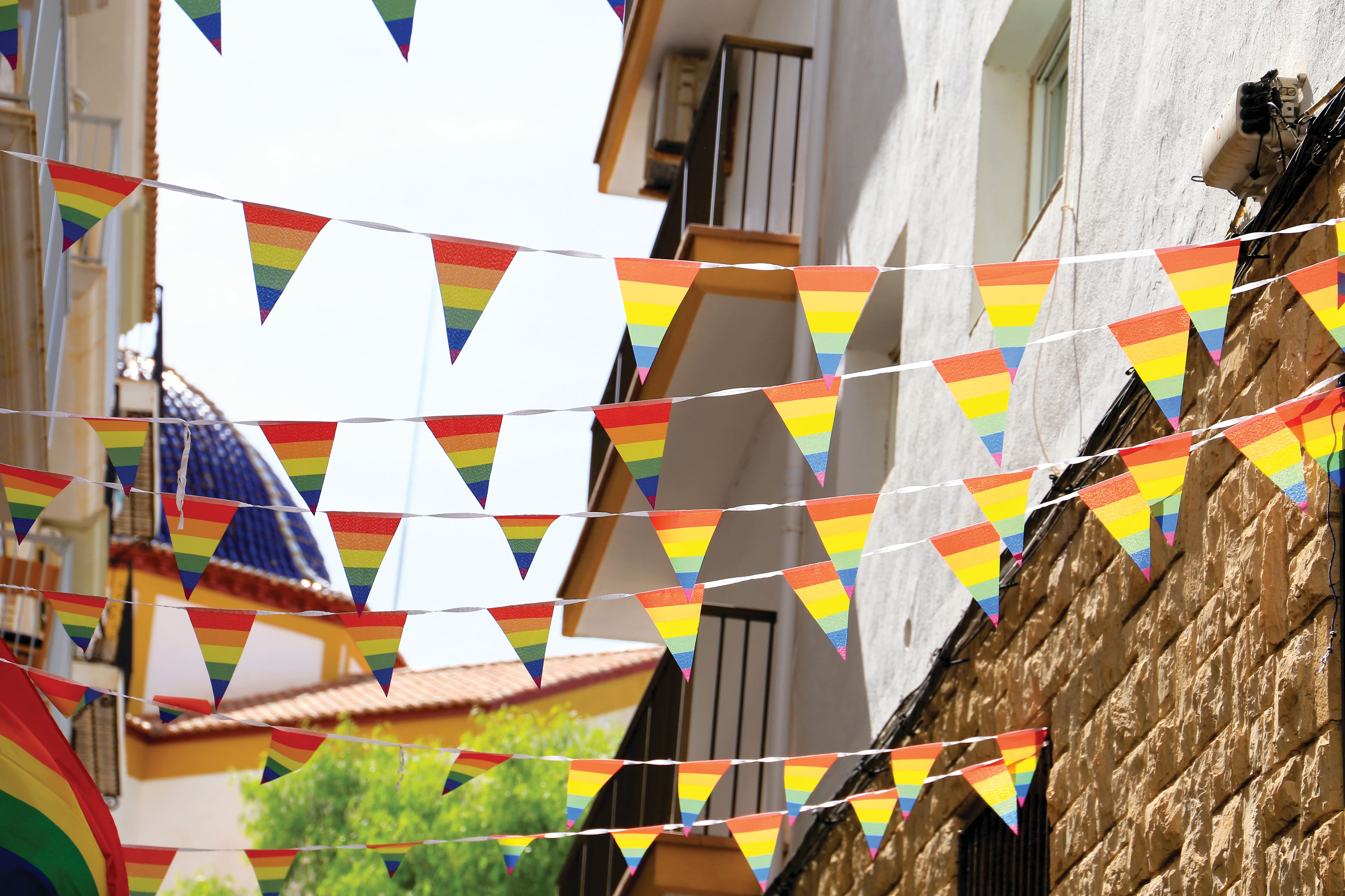 Colourful rainbow bunting hangs across a narrow street lined with traditional buildings and balconies. The festive decorations stretch between the buildings in bright sunshine, creating a vibrant atmosphere associated with Pride celebrations.