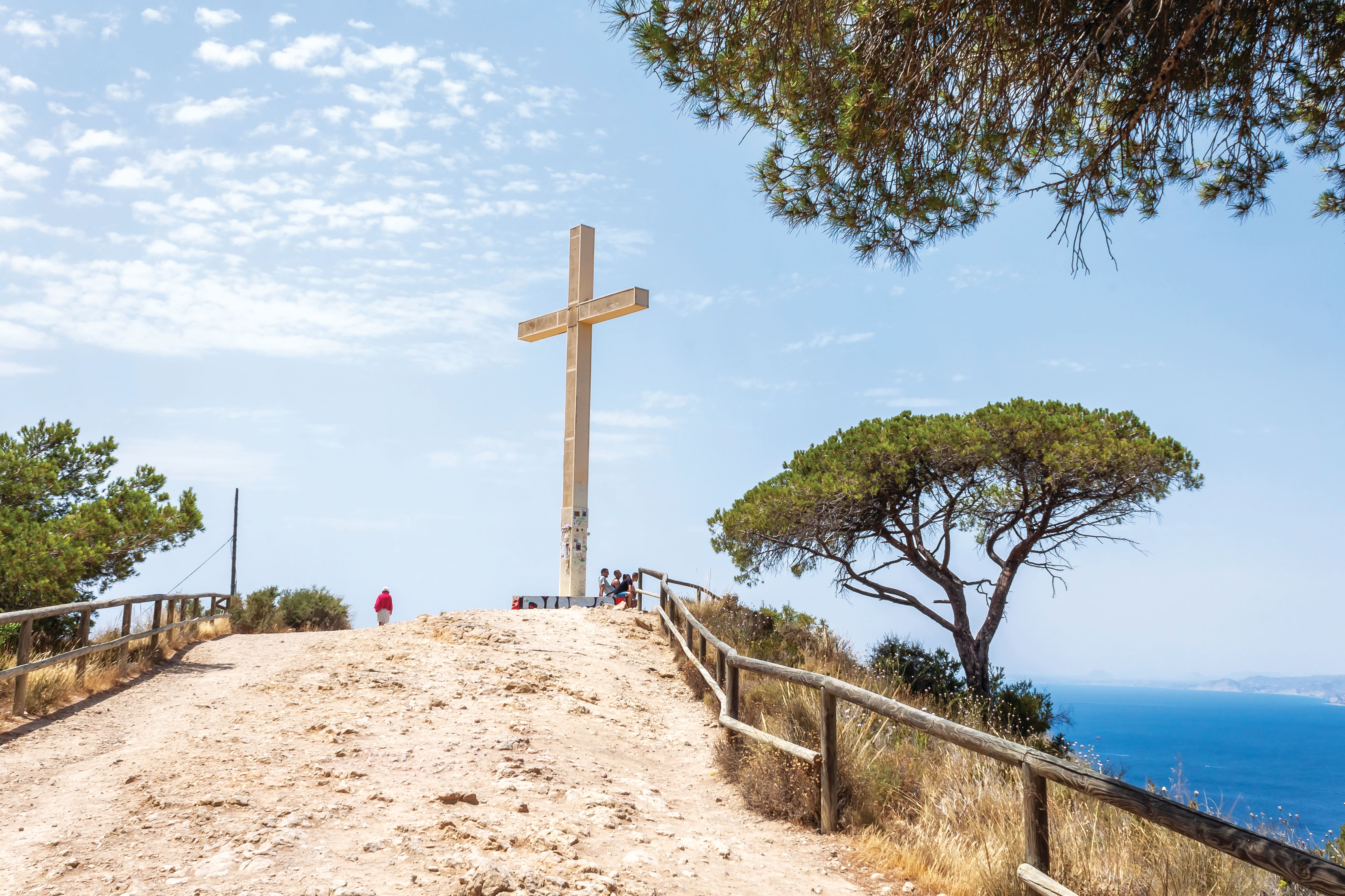 The Benidorm Cross stands at the top of a rocky hillside, reached by a dirt path lined with wooden fencing. Framed by pine trees and set against a bright blue sky with scattered clouds, the viewpoint overlooks the Mediterranean coastline below.