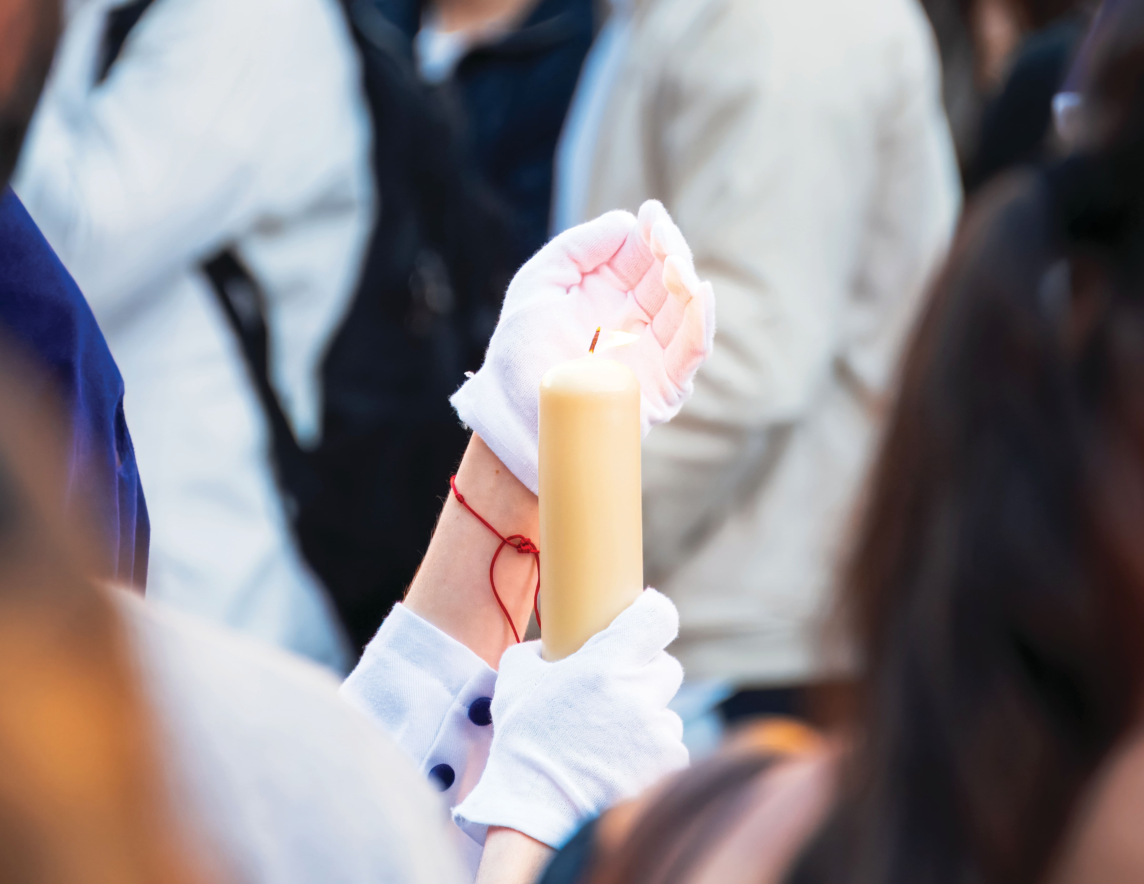 Close-up of a candlelight procession, with a person wearing white gloves holding a large lit candle among a crowd. The background is softly blurred, drawing attention to the glowing flame and ceremonial atmosphere.