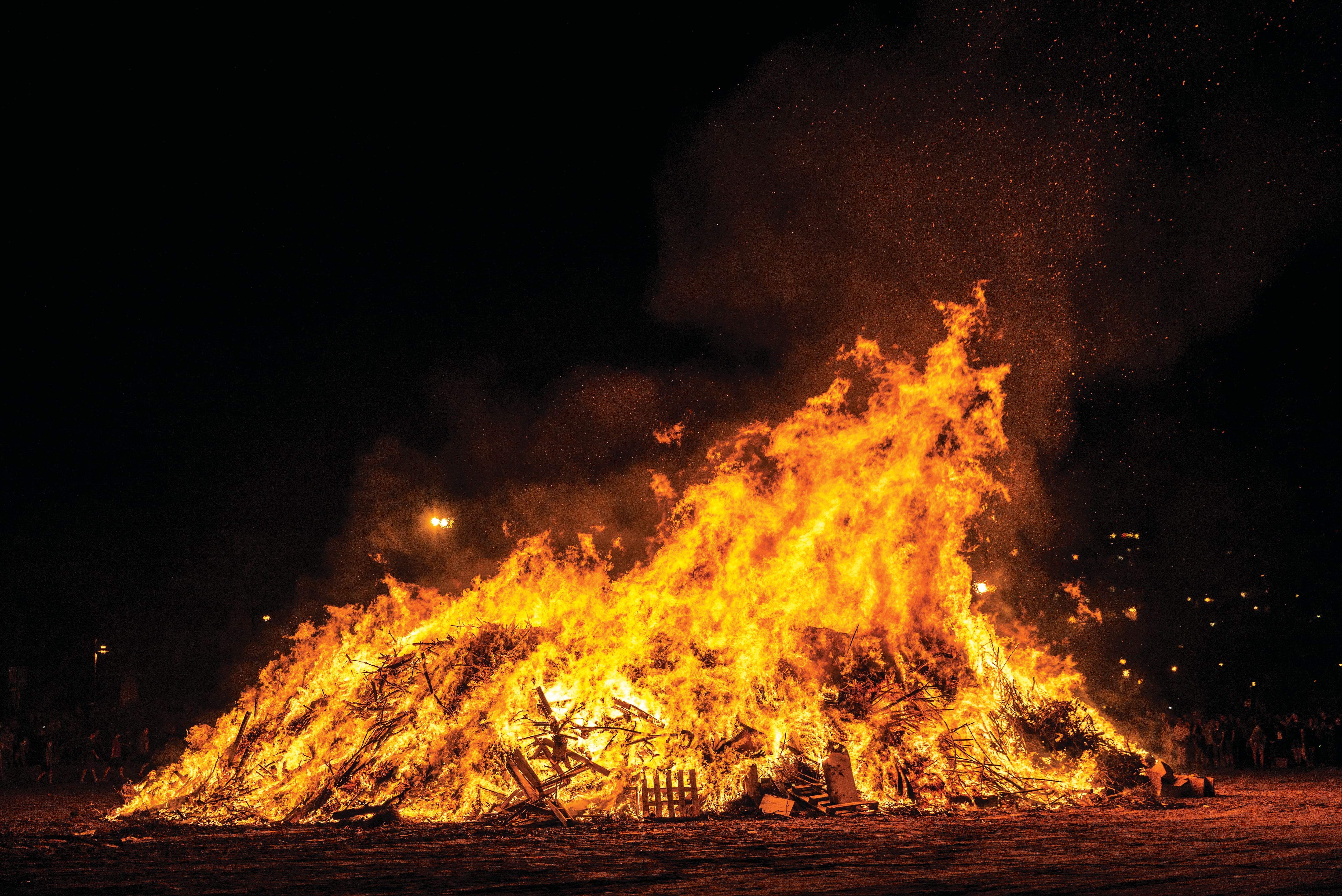 Large bonfire blazing at night, with towering flames and glowing embers rising into the dark sky. The fire is built from stacked wood and debris, creating a dramatic display that illuminates the surrounding open space.