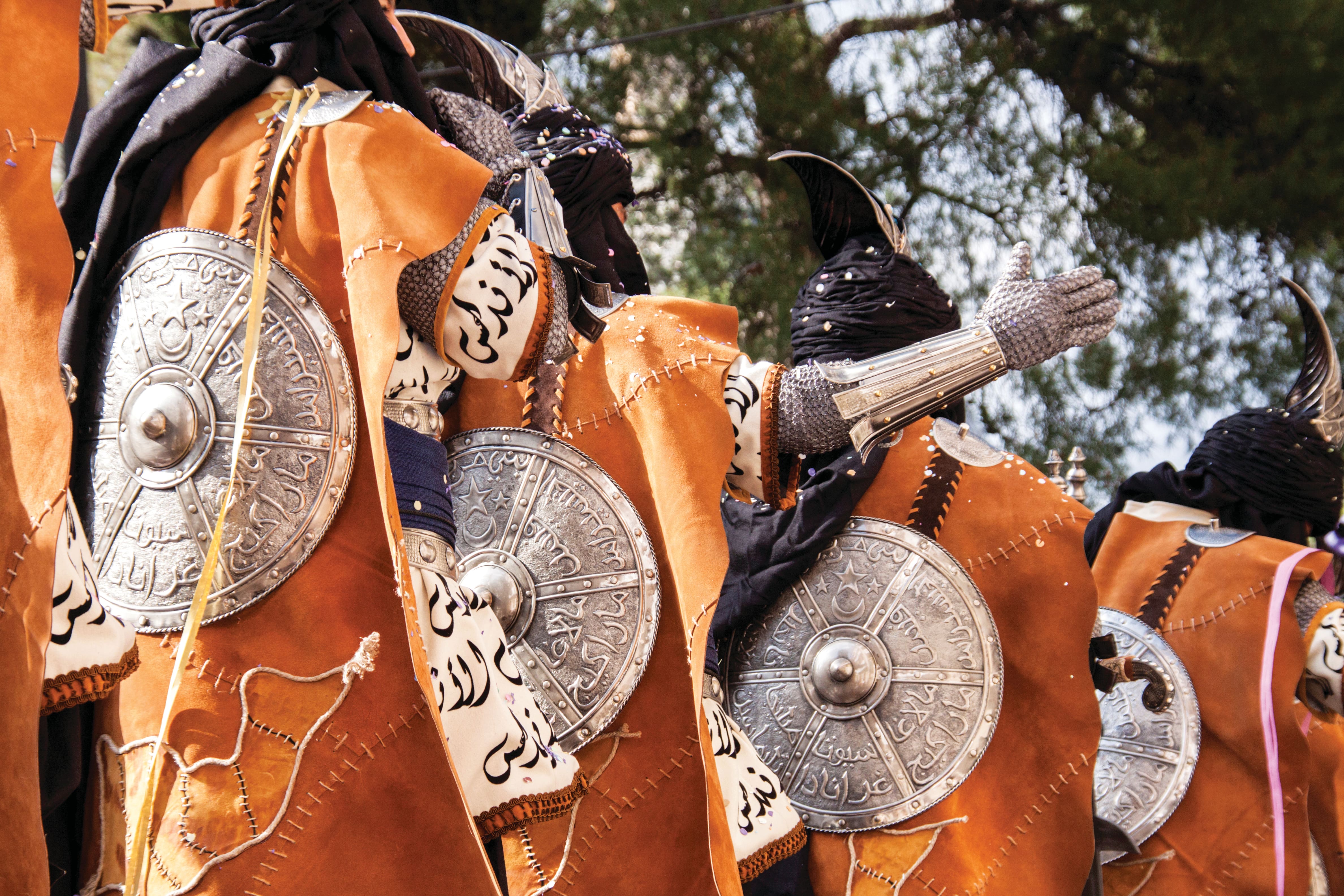 Participants in traditional Moorish-inspired costumes parade in formation, wearing orange robes, chainmail-style armour, ornate shields and curved black headpieces. The detailed outfits and ceremonial attire are part of a colourful Moors and Christians.