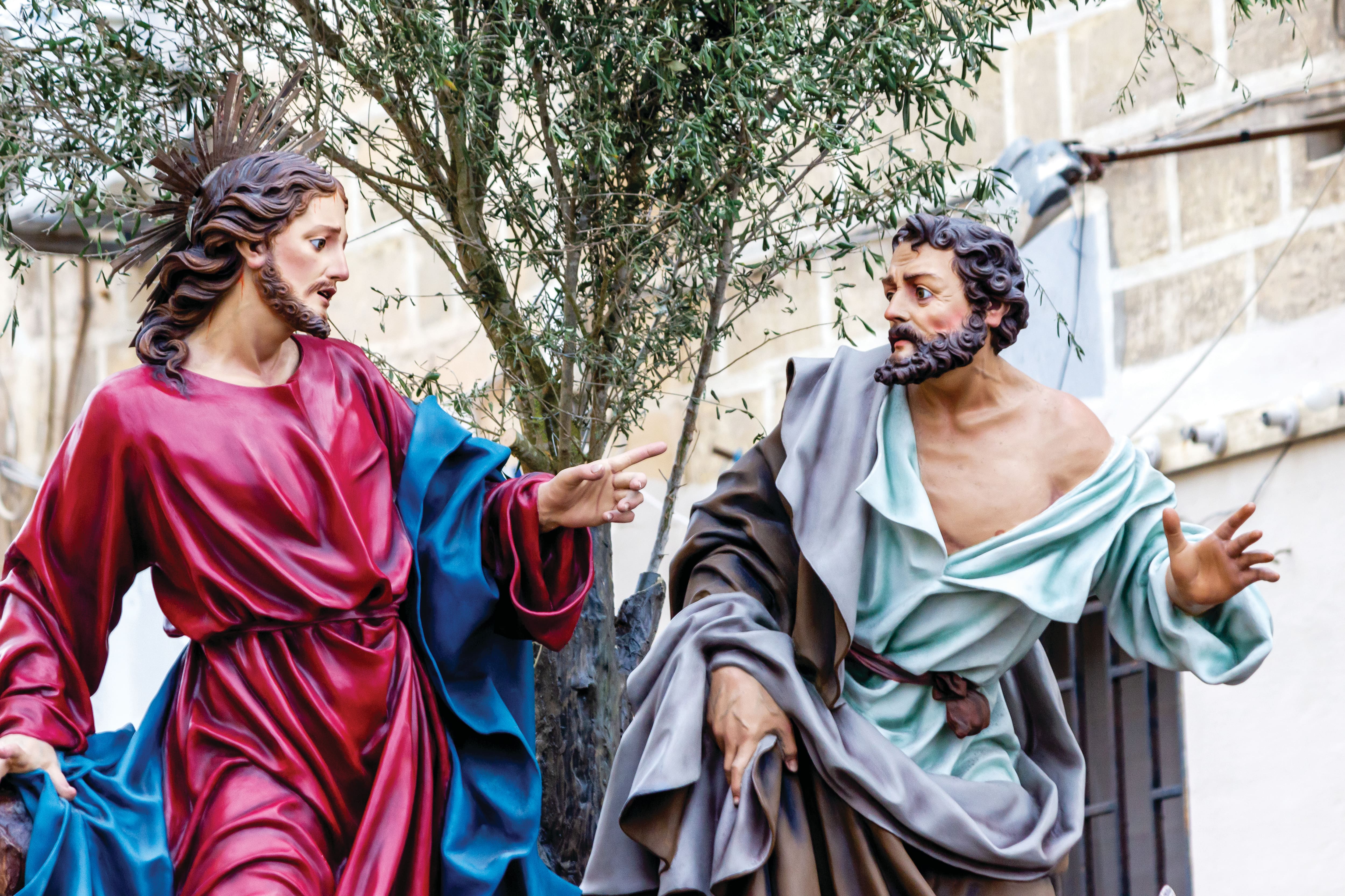 Religious procession statue depicting two biblical figures in flowing robes, one pointing towards the other, displayed outdoors beneath an olive tree. The detailed sculptures are part of a traditional Holy Week celebration