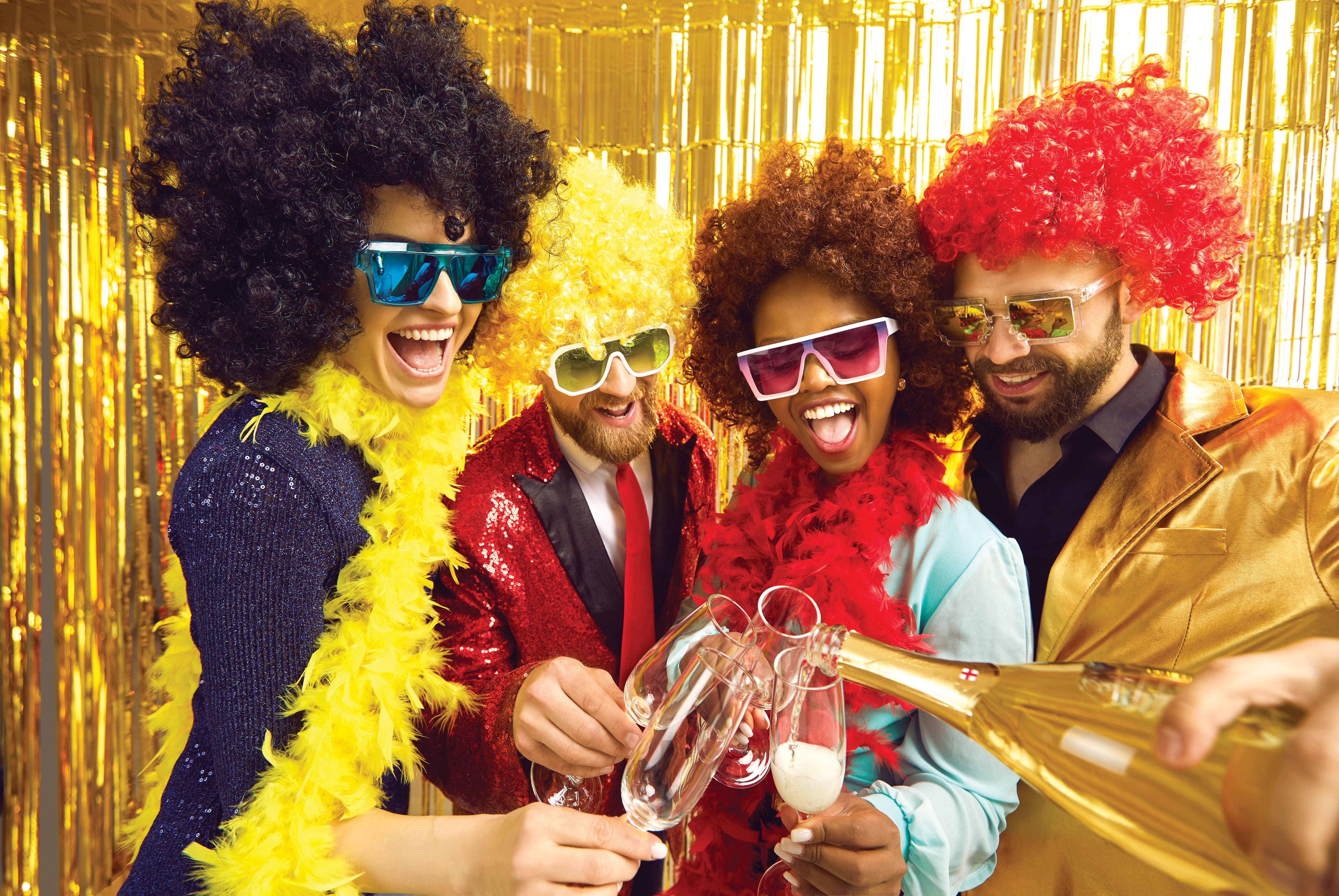 Group of partygoers dressed in colourful fancy dress costumes, including bright curly wigs, oversized sunglasses and feather boas, as sparkling wine is poured into glasses. A gold shimmering backdrop adds to the festive celebration atmosphere.