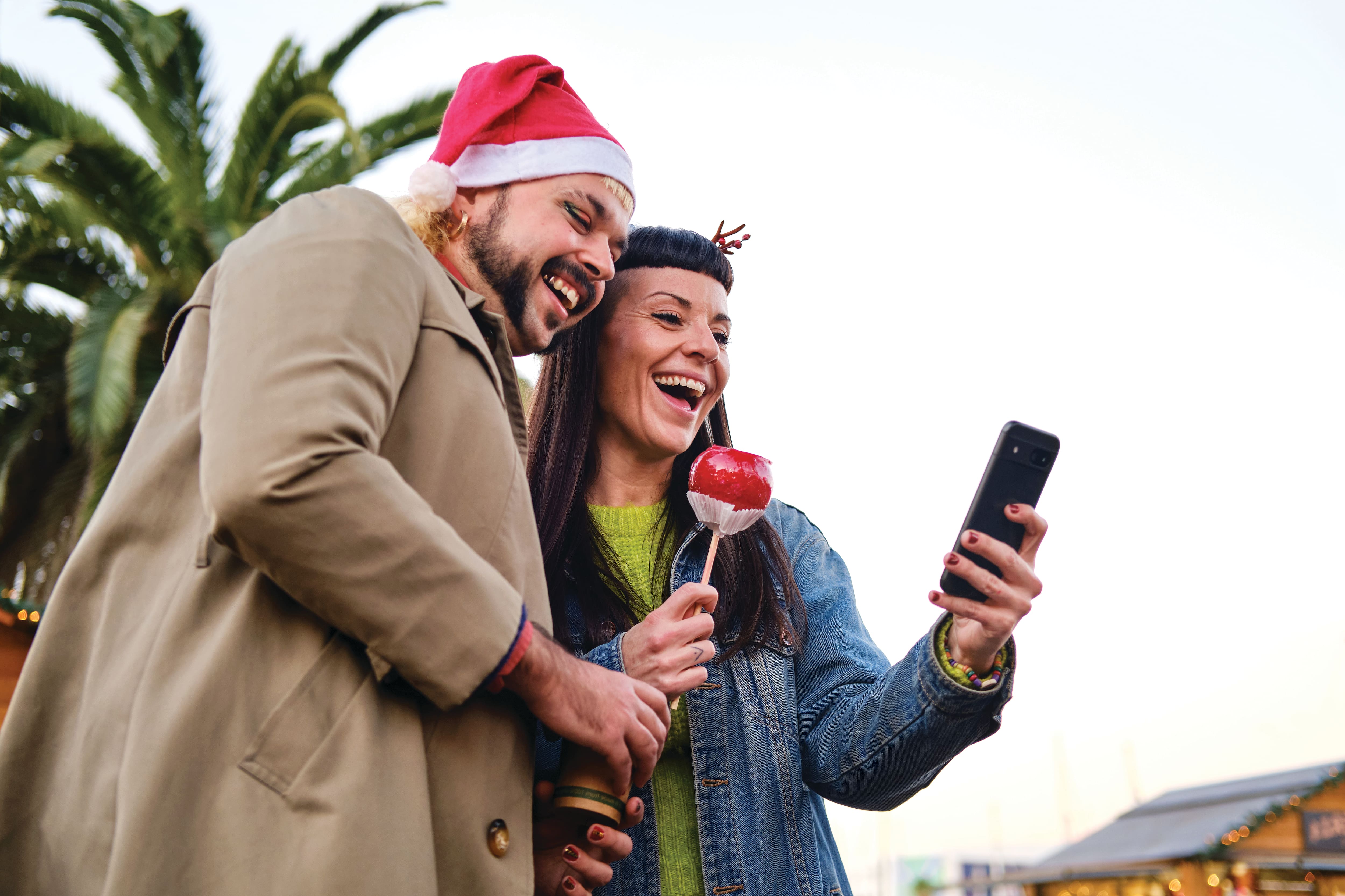 Two people wearing festive Christmas accessories, including a Santa hat and reindeer antlers, look at a smartphone while standing outdoors near palm trees. One person holds a red candy apple, creating a cheerful holiday scene in a warm-weather setting.