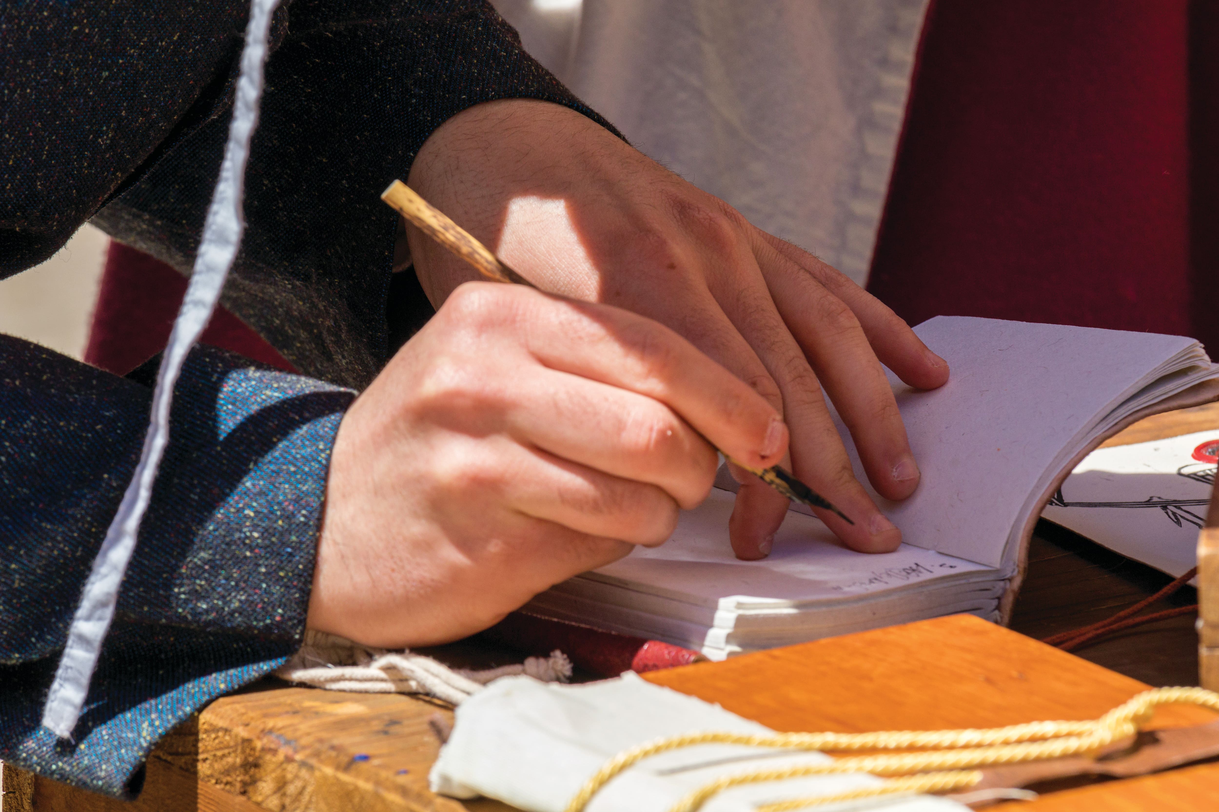 Close-up of a person writing in a notebook with a traditional dip pen at a wooden table. The scene features period-style clothing, handwritten pages and historical props, suggesting a historical re-enactment or cultural demonstration.