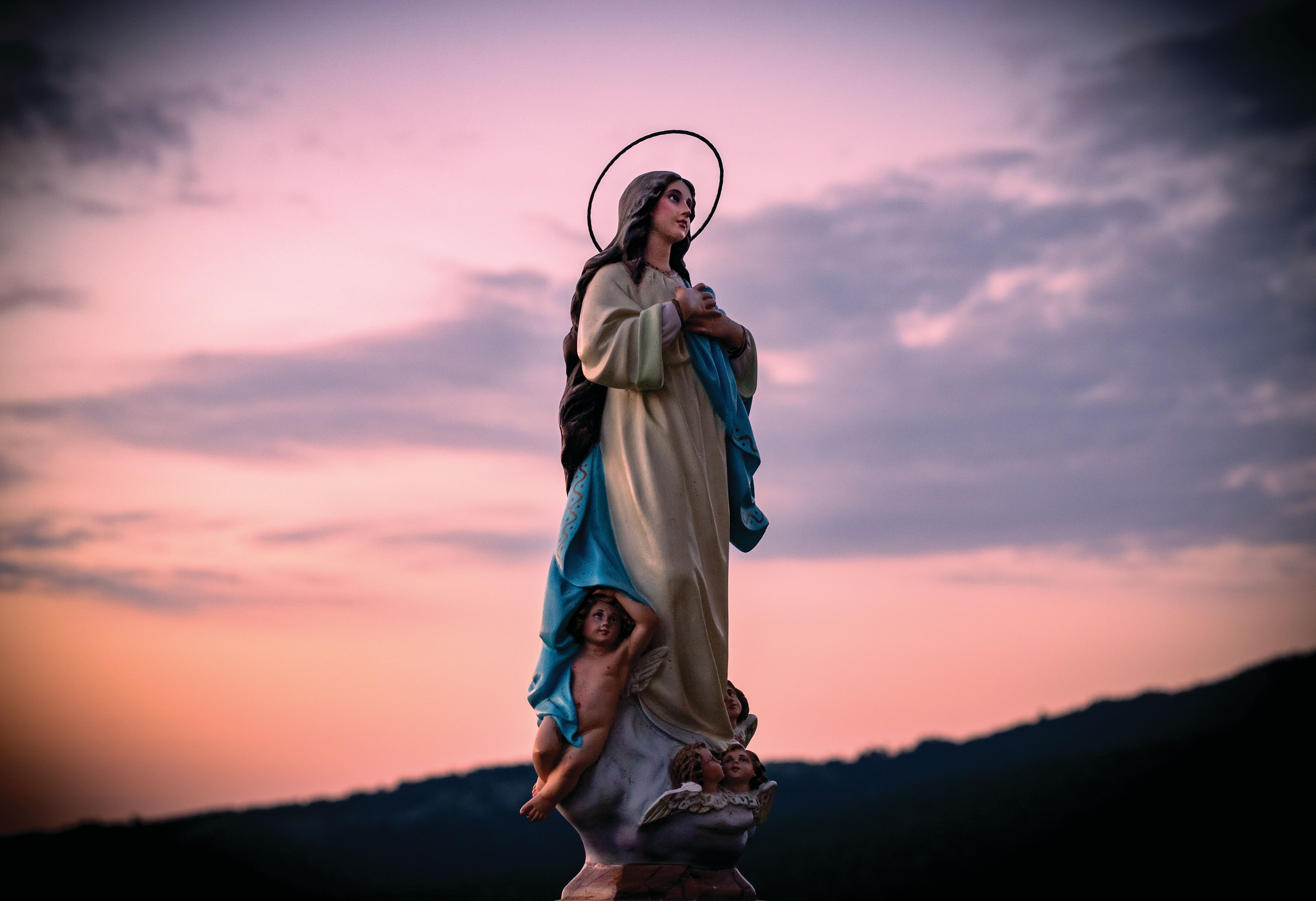 Statue of the Virgin Mary standing on a pedestal surrounded by cherubs, with a halo above her head and a blue robe draped over a cream-coloured gown. The religious figure is silhouetted against a pink and purple sunset sky with hills in the background