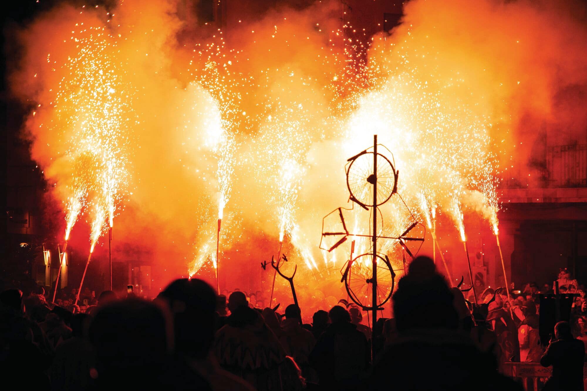 A crowd of people silhouetted in the foreground watches a dramatic outdoor fireworks display