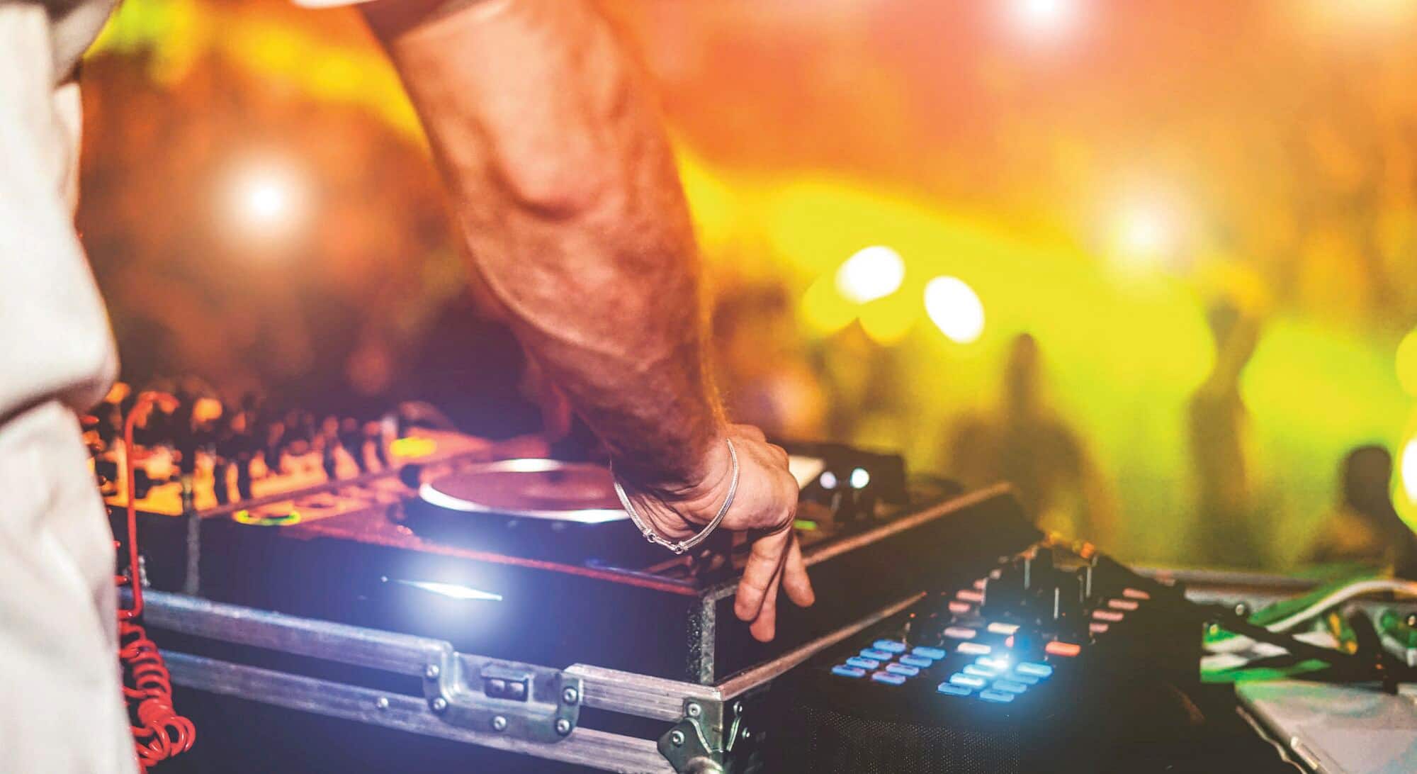 An arm of a DJ mixing music on a turntable console at a party with bright lights and a blurred crowd in the background