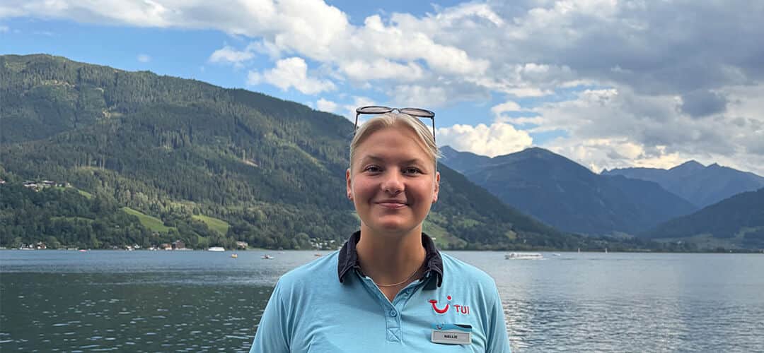 A woman with blonde hair faces the camera with a lake behind. 