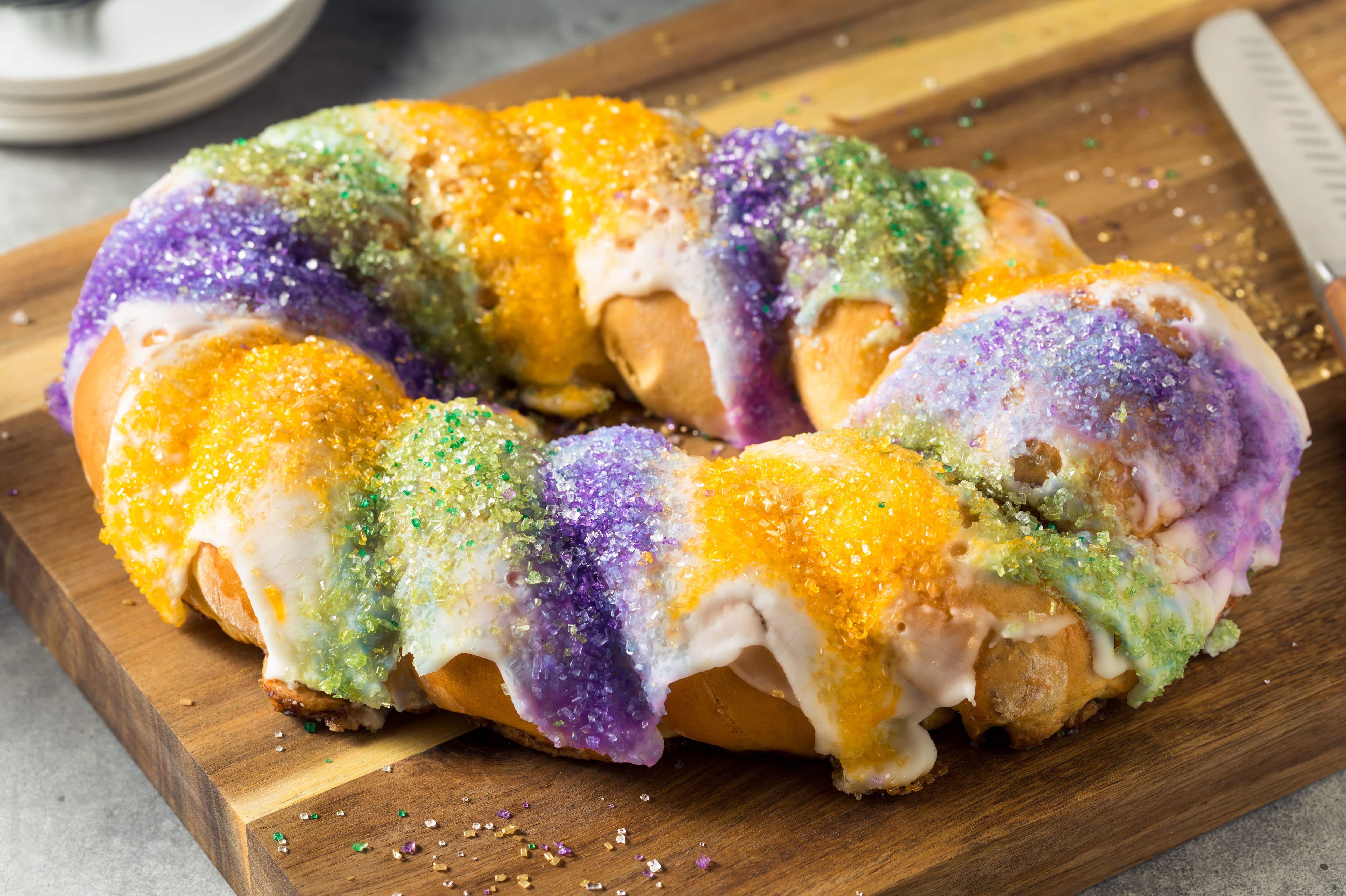 A ring-shaped cake topped with white icing and purple, green and gold sugar crystals sits on a wooden serving board.