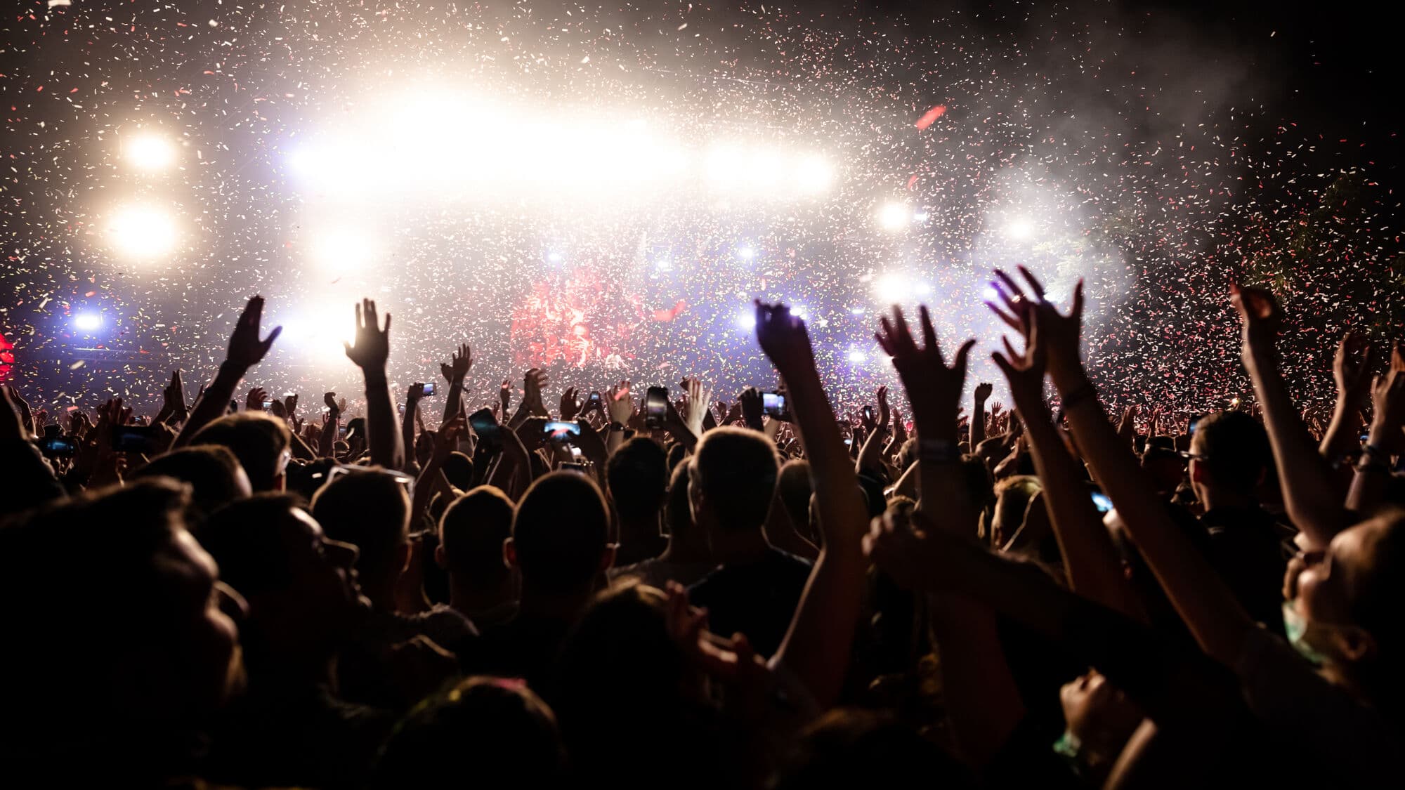 Crowd gathered at an outdoor concert with raised hands, stage lights and confetti in the air.