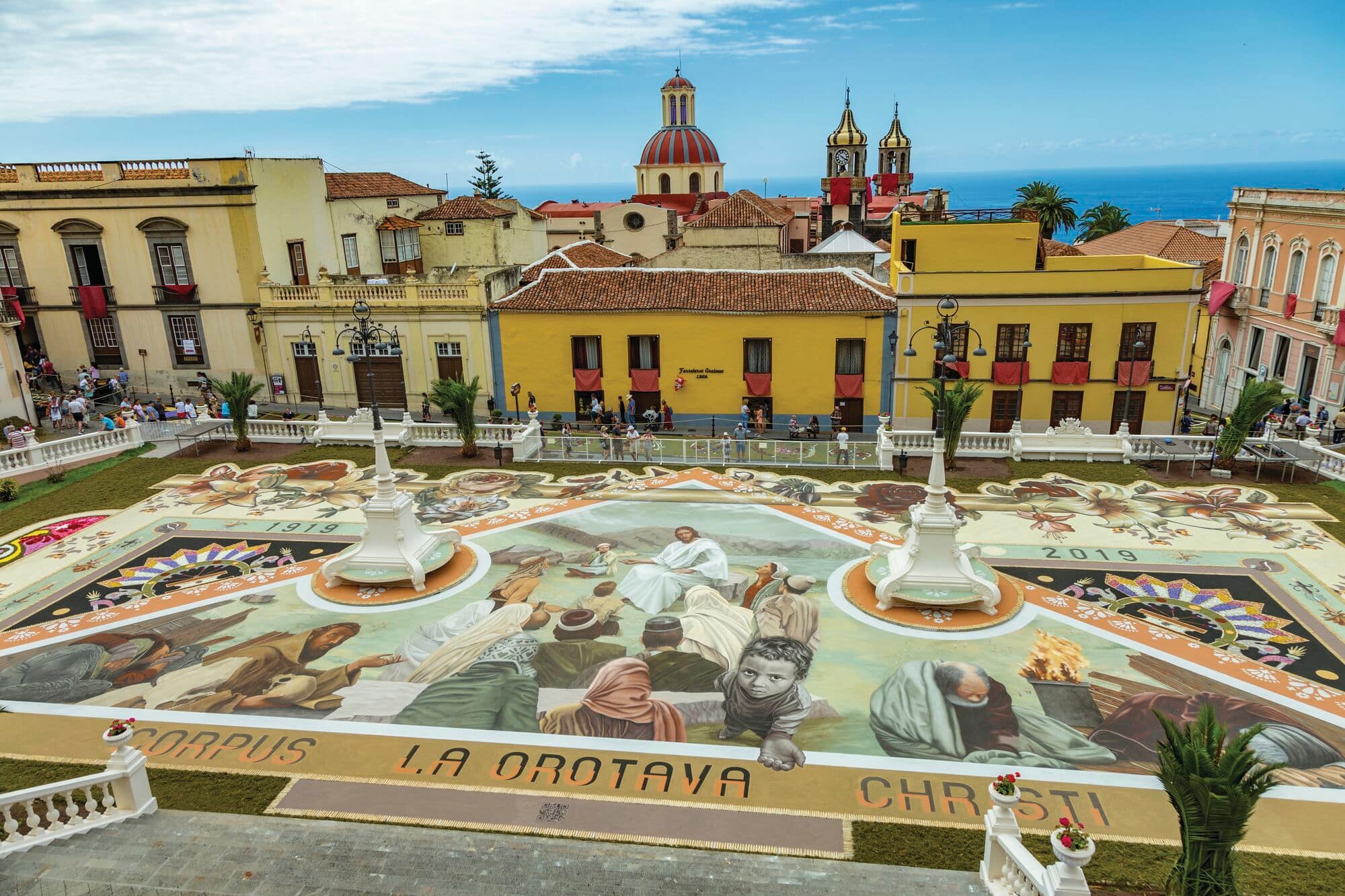 Large colourful flower mural in a historic town square, depicting religious and cultural scenes. Surrounded by yellow and pastel-coloured buildings, church domes and towers in the background, and the blue ocean visible on the horizon under a clear sky.