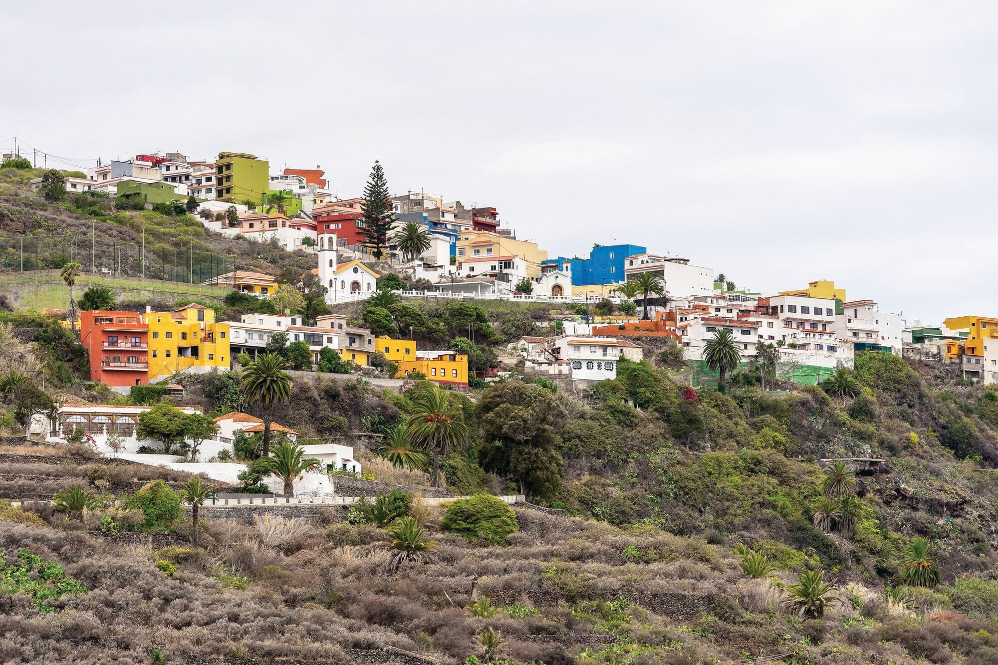 Colourful hillside village overlooking a rugged landscape, with bright houses in shades of yellow, blue, orange and white scattered across the slopes. Palm trees and terraced vegetation add to the scenic rural setting.