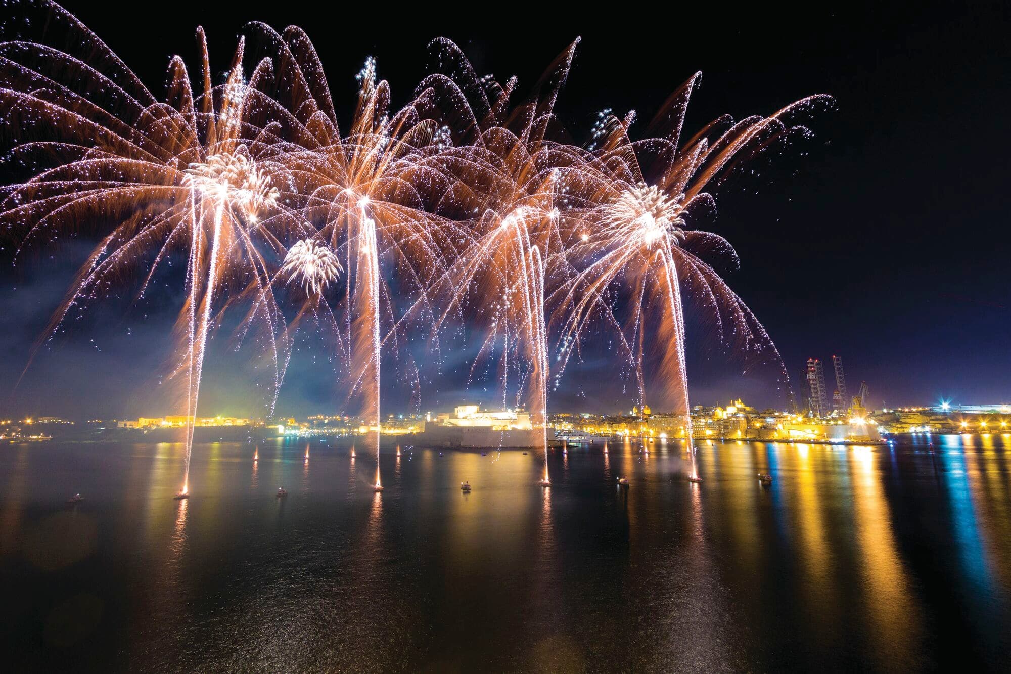 Golden fireworks cascade over a calm harbour at night, reflecting across the water. Bright bursts illuminate the shoreline and marina lights, creating a spectacular display against the dark sky.