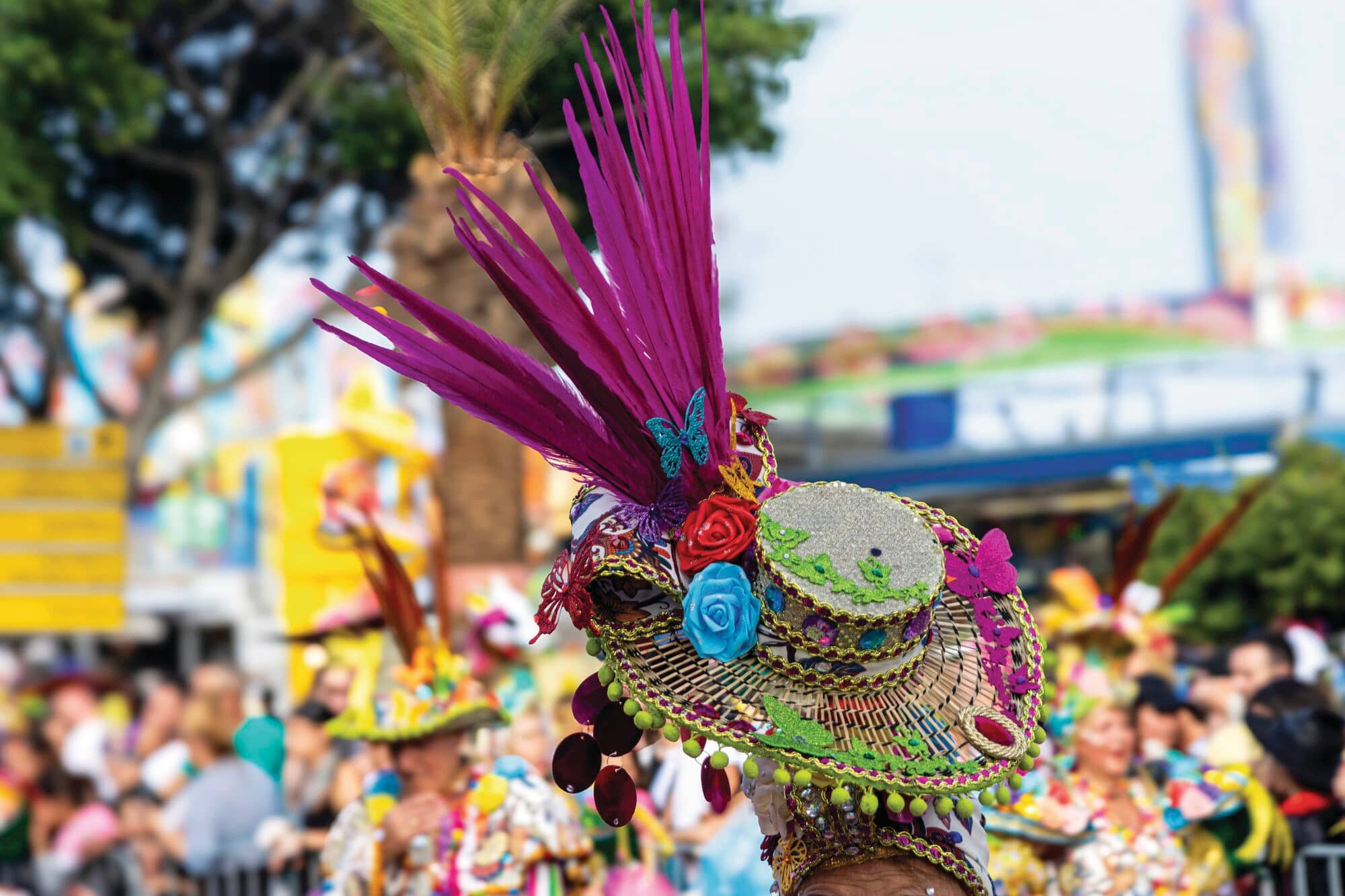 Image of woman wearing colourful hat with large bright feathers with blurred carnival scene in the background.
