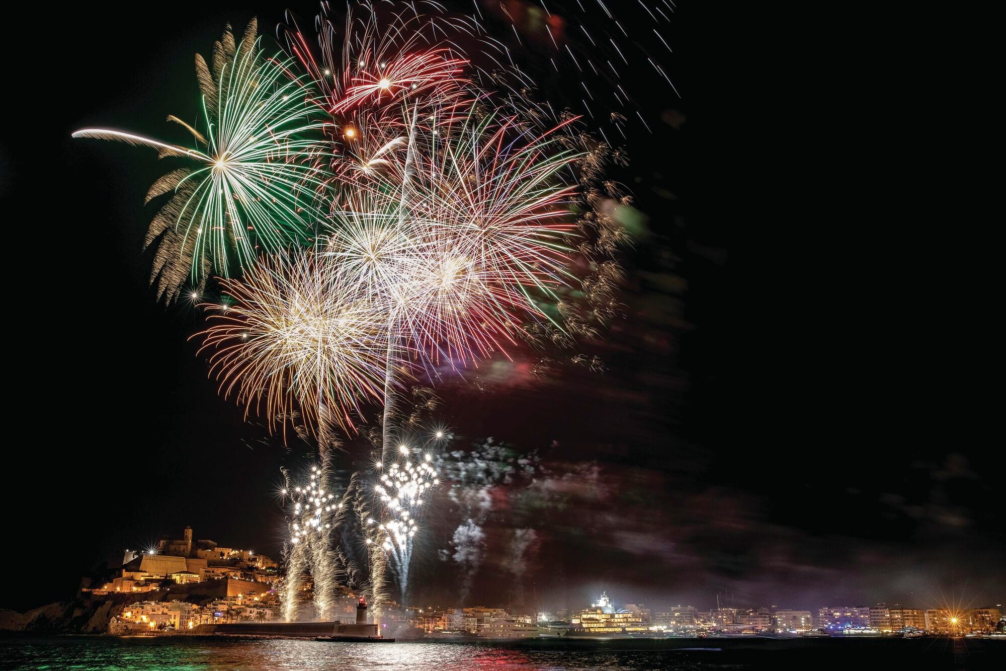 Colourful fireworks burst across the night sky above a coastal town, lighting up the waterfront and reflecting on the sea. Bright displays of red, green and gold create a festive atmosphere against the dark backdrop.  Provide your feedback on BizChat