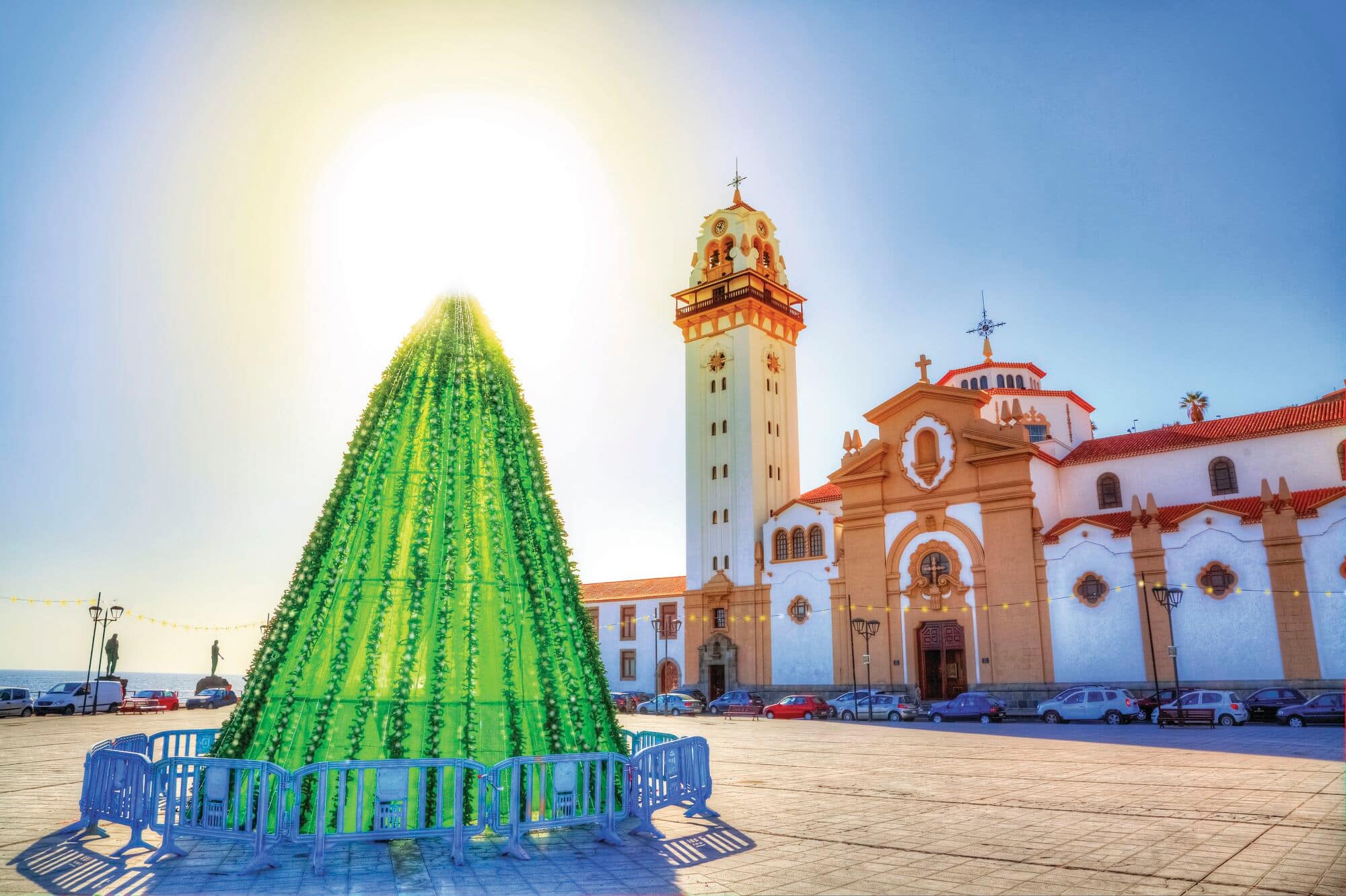 Large green Christmas tree decoration stands in a sunlit town square beside a historic church with a tall bell tower. The bright blue sky and traditional architecture create a festive and picturesque setting.