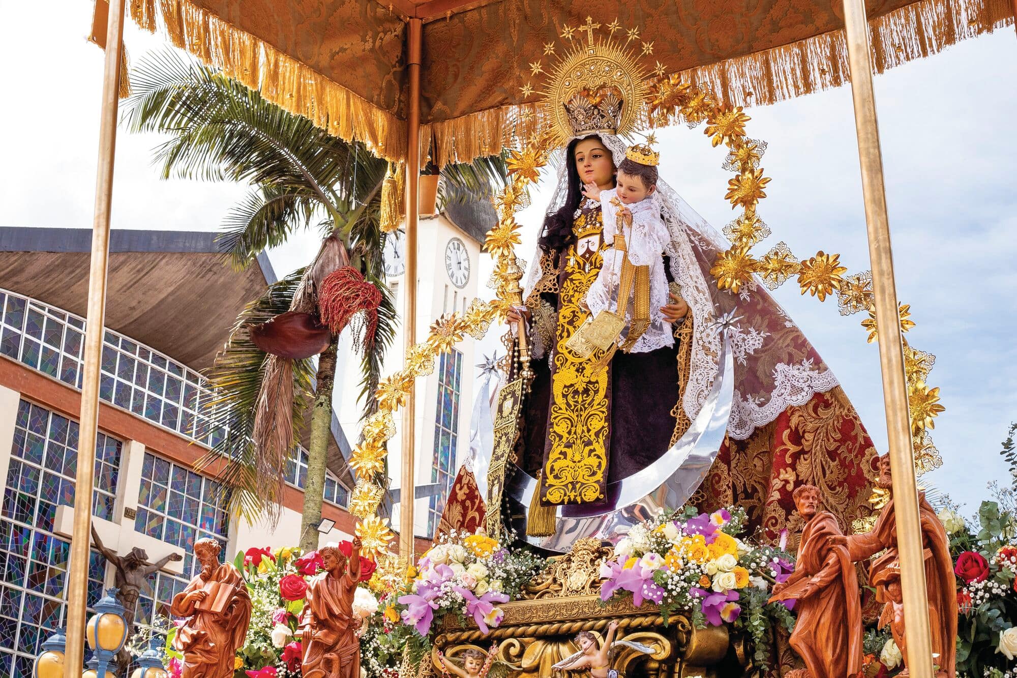 Decorated religious statue displayed on a flower-filled platform during a cultural procession, framed by a gold-trimmed canopy. Palm trees, a clock tower and buildings can be seen in the background beneath a bright sky.