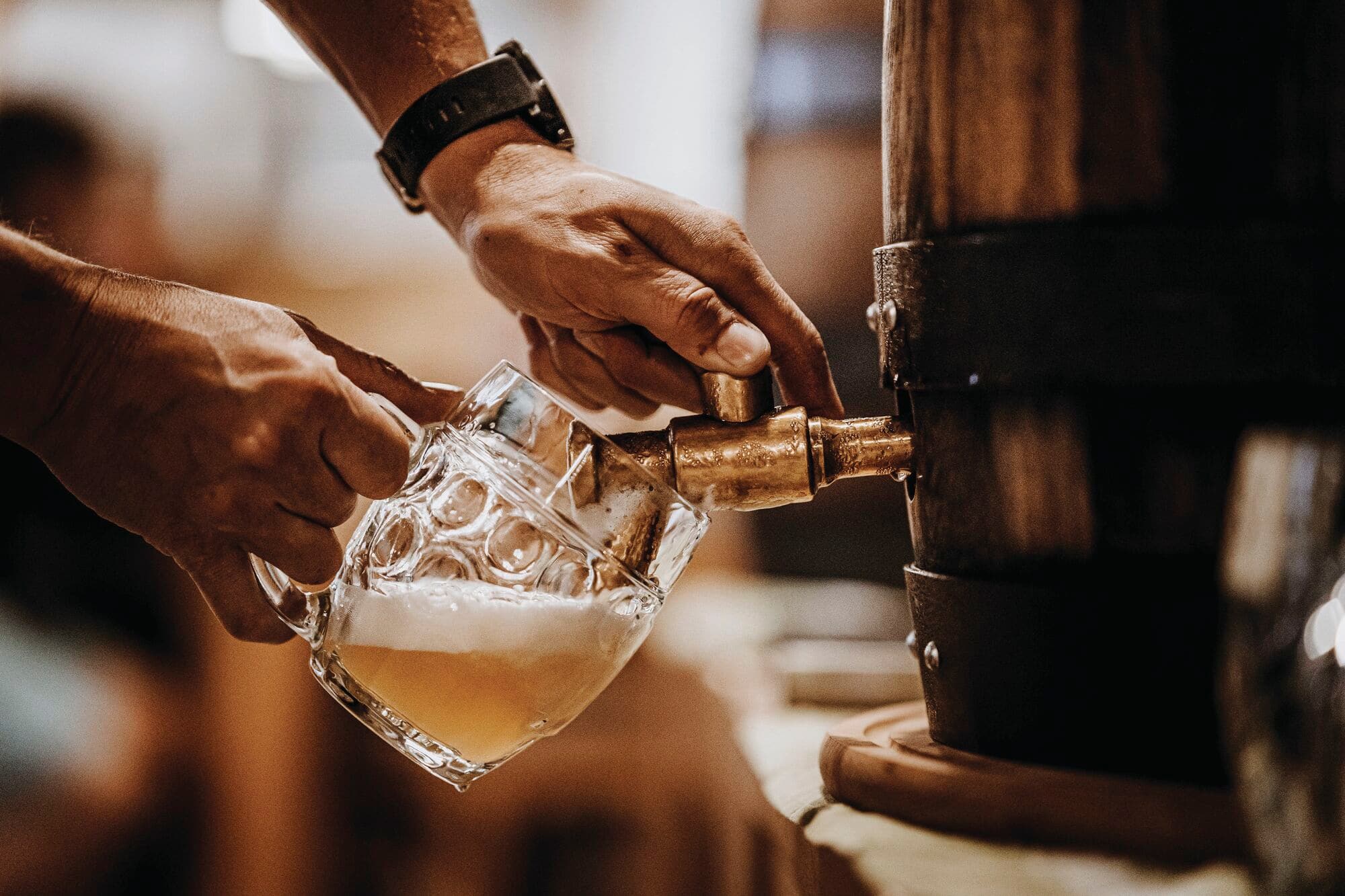 A person pouring a golden beer, from a small wooden tabletop keg, into a glass beer mug