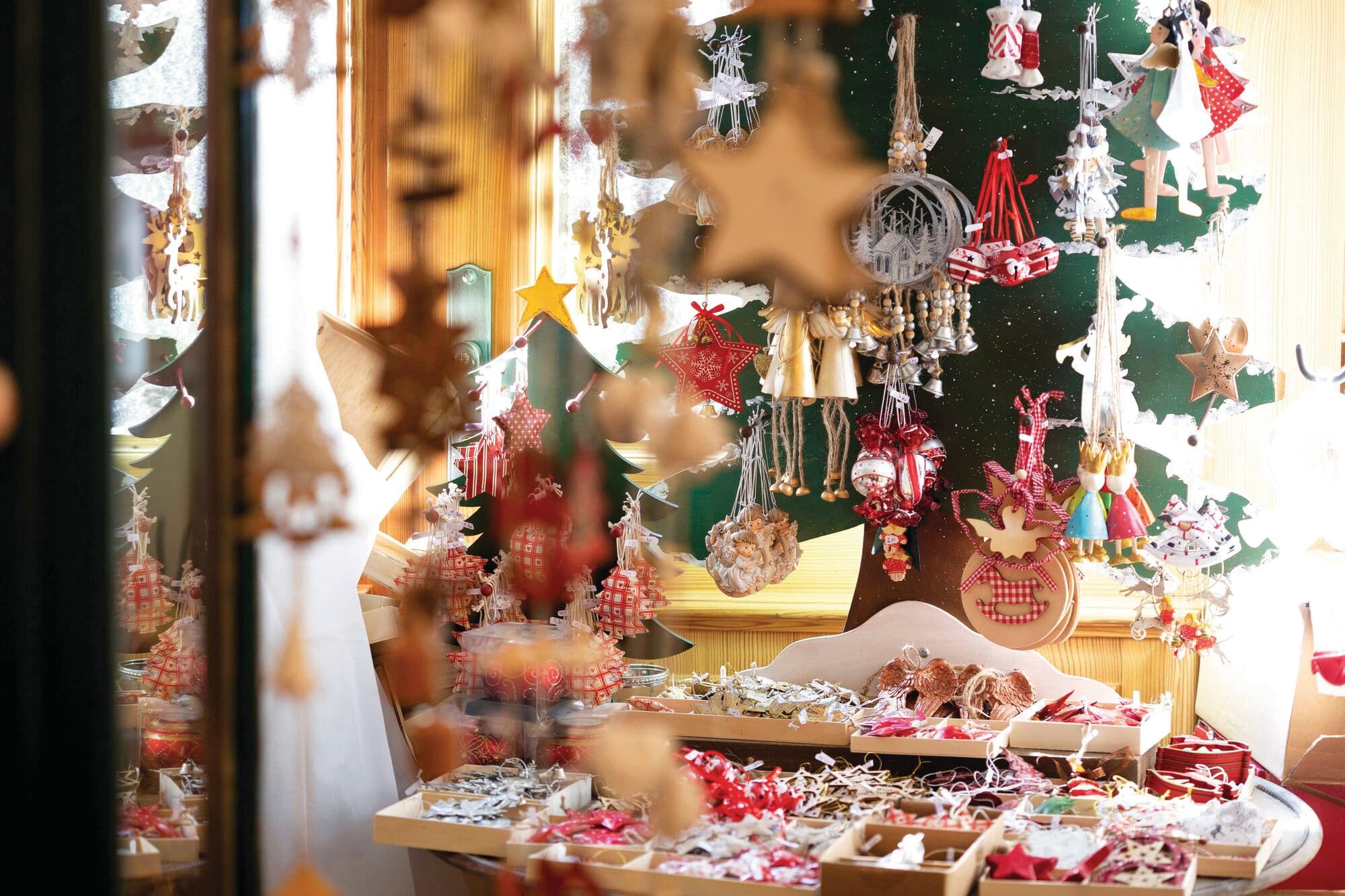 A table filled with small wooden trays displaying various holiday items, with star and bell decorations hanging from the ceiling in the foreground and a large green Christmas tree decoration the background