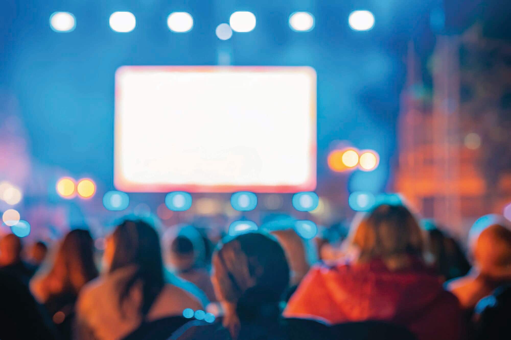 An audience sitting outdoors at night, facing a large lighted screen during an open-air movie screening