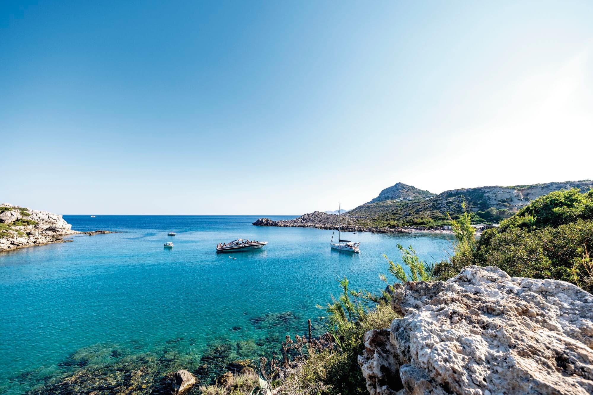 Scenic Mediterranean cove with crystal-clear turquoise water, three anchored boats, white limestone cliffs, and green hillsides under a bright blue sky.